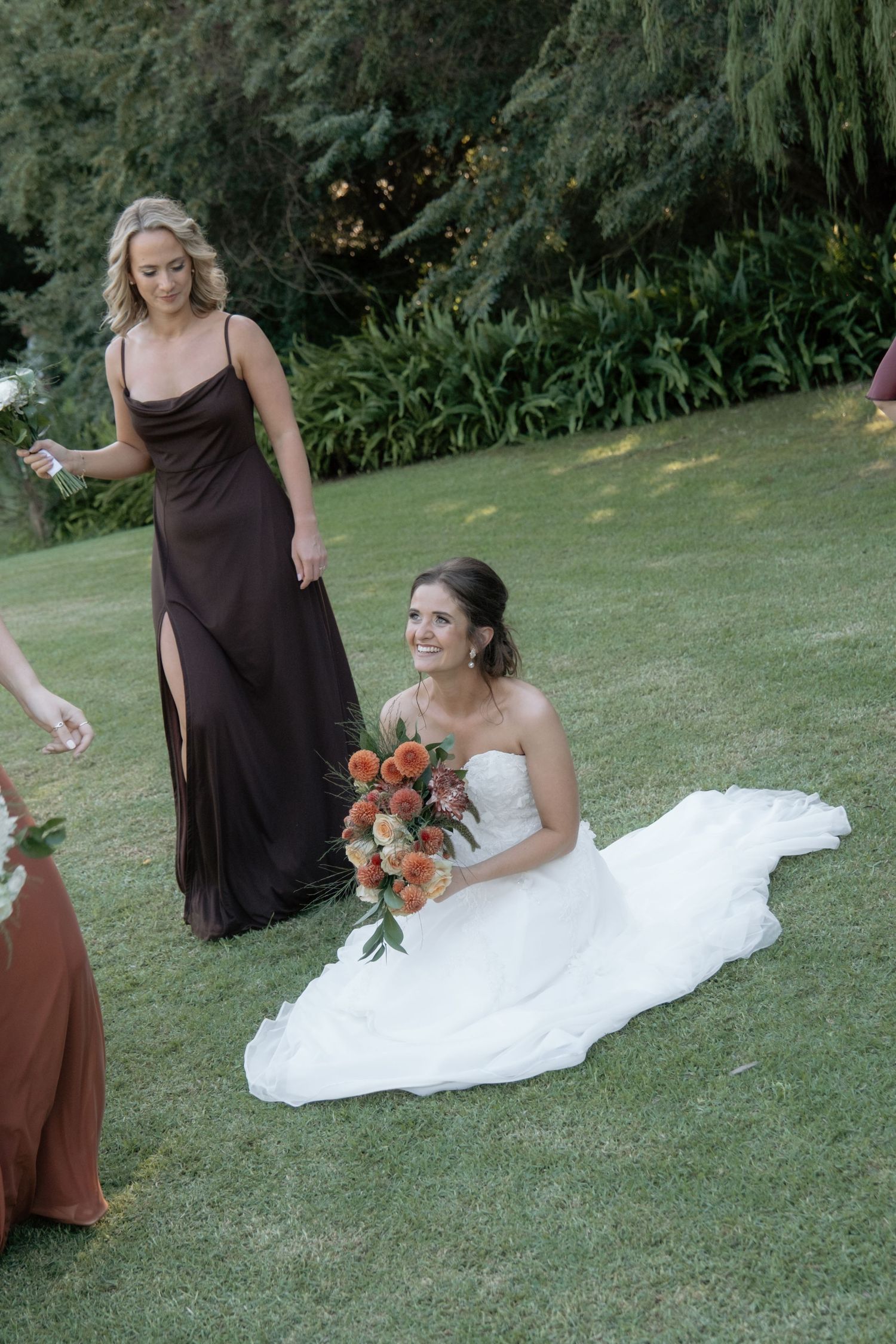 Bride in white gown sitting on grass holding orange bouquet with bridesmaid in brown dress nearby.