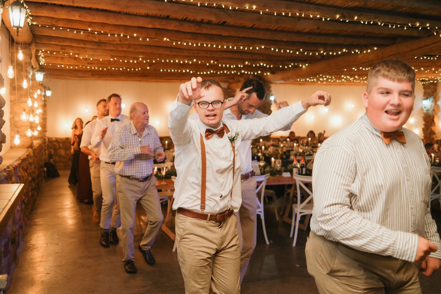 Groomsmen and guests dance joyfully at a rustic wedding reception decorated with string lights.