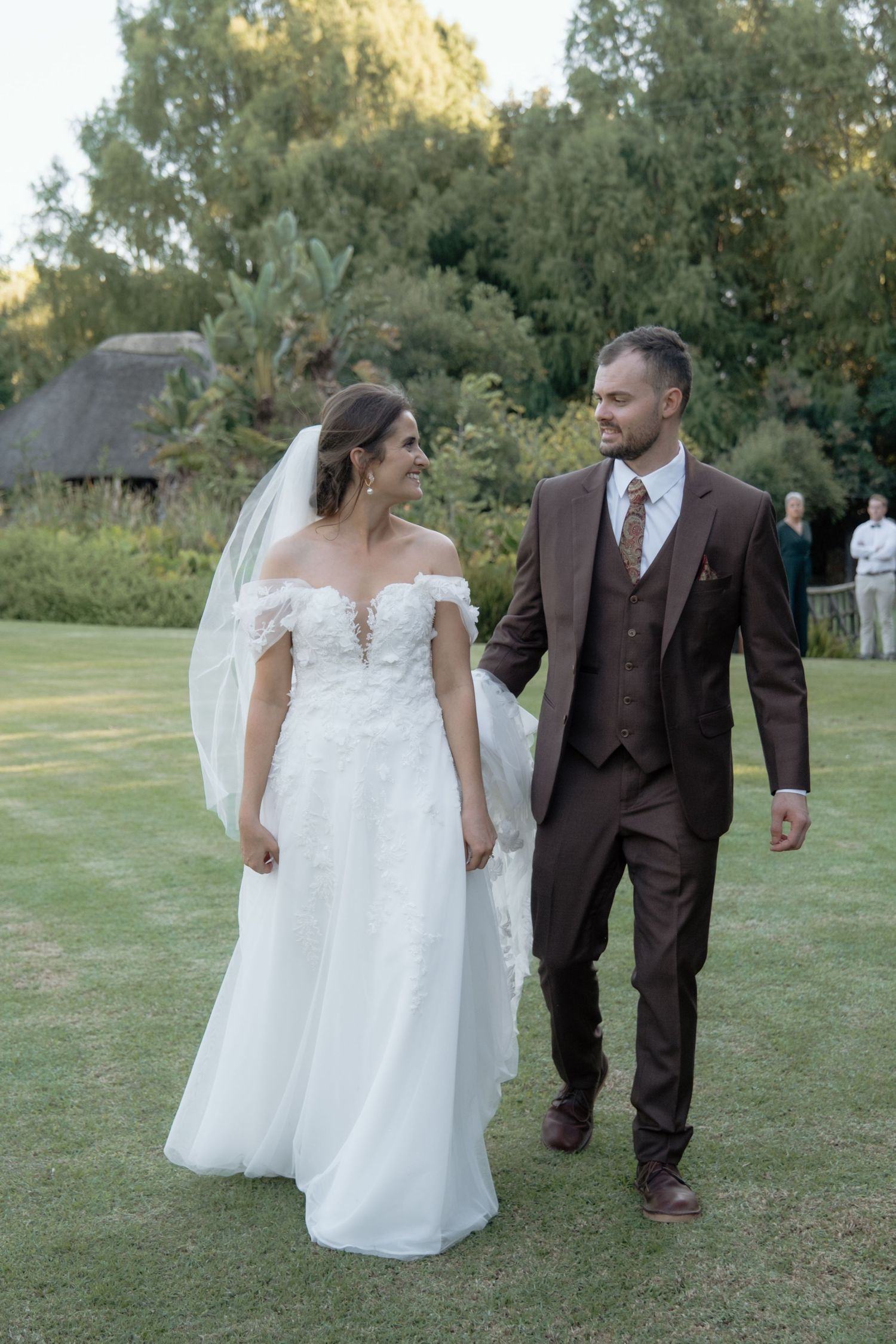 Bride and groom walk hand in hand across green lawn smiling at each other after wedding.