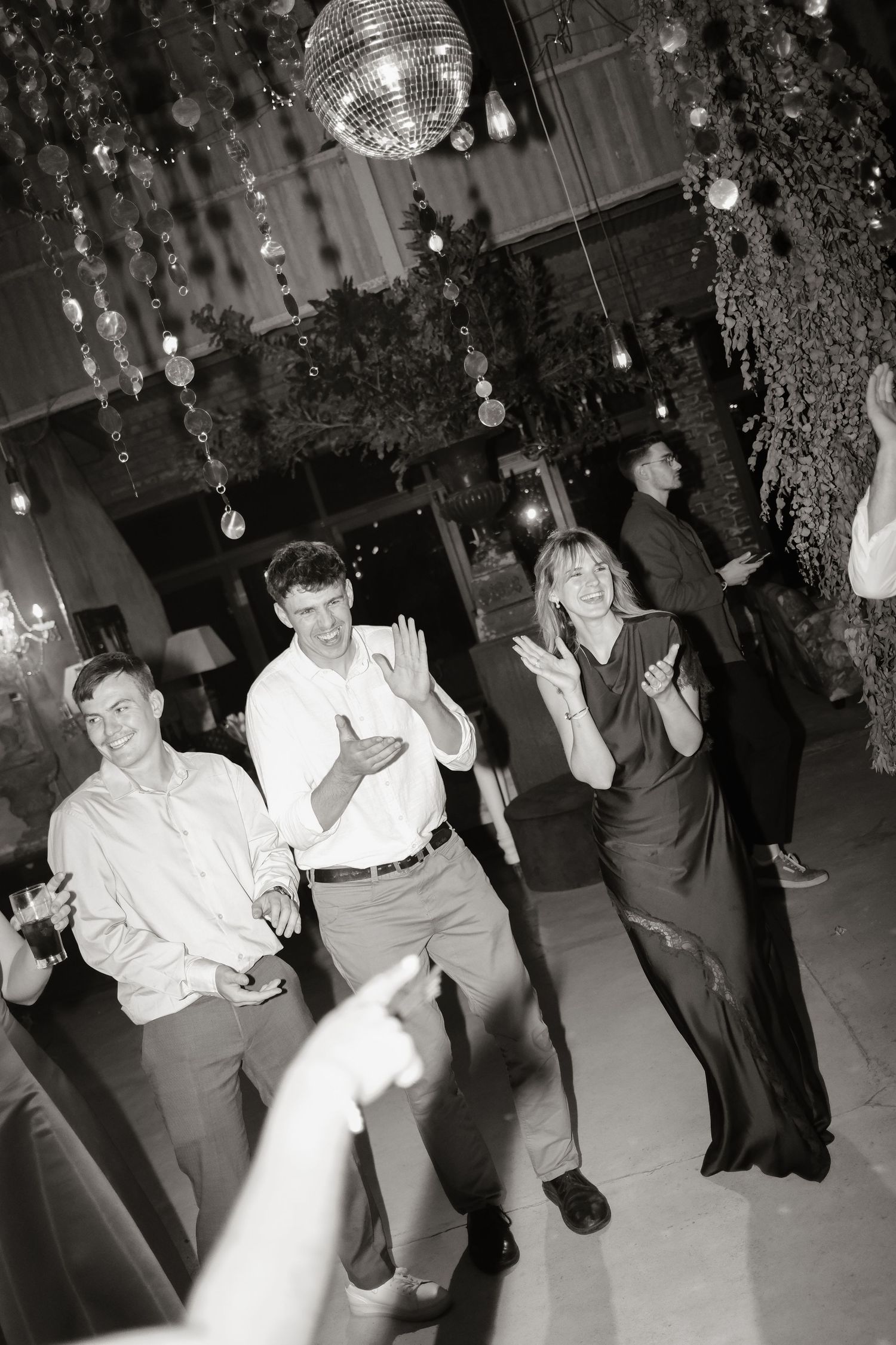 Black and white photo of wedding guests dancing and clapping under a disco ball at a festive reception.