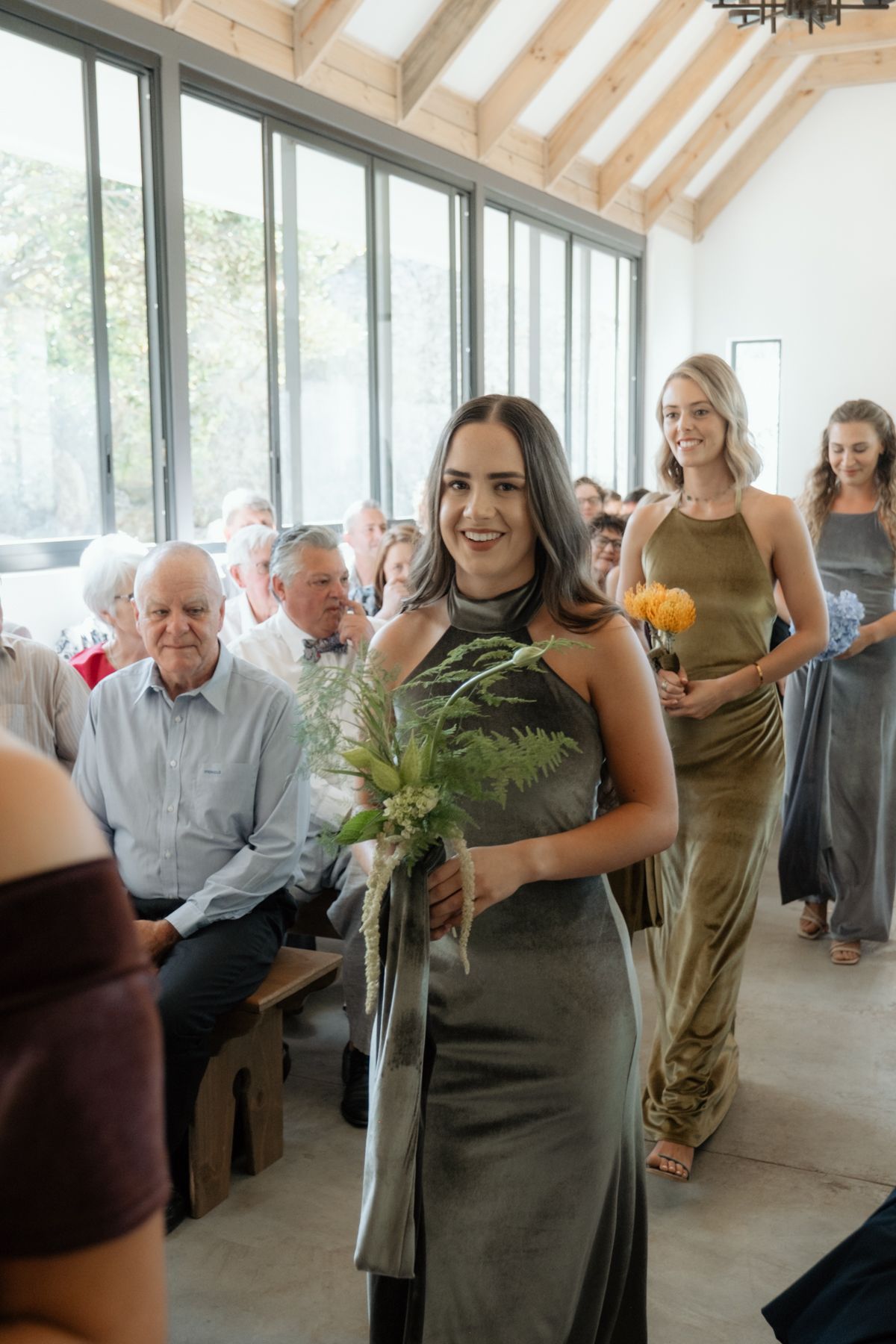 Bridesmaids in olive velvet dresses walk down the aisle at an elegant wedding ceremony with guests seated on wooden pews.
