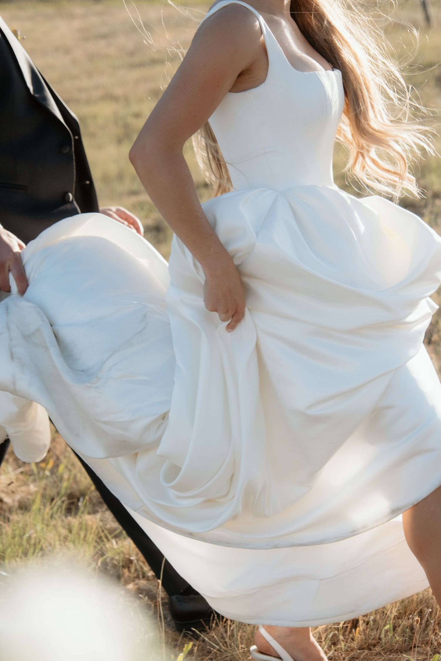 Bride in a flowing white ball gown walking outdoors with groom in a sunlit field on wedding day.