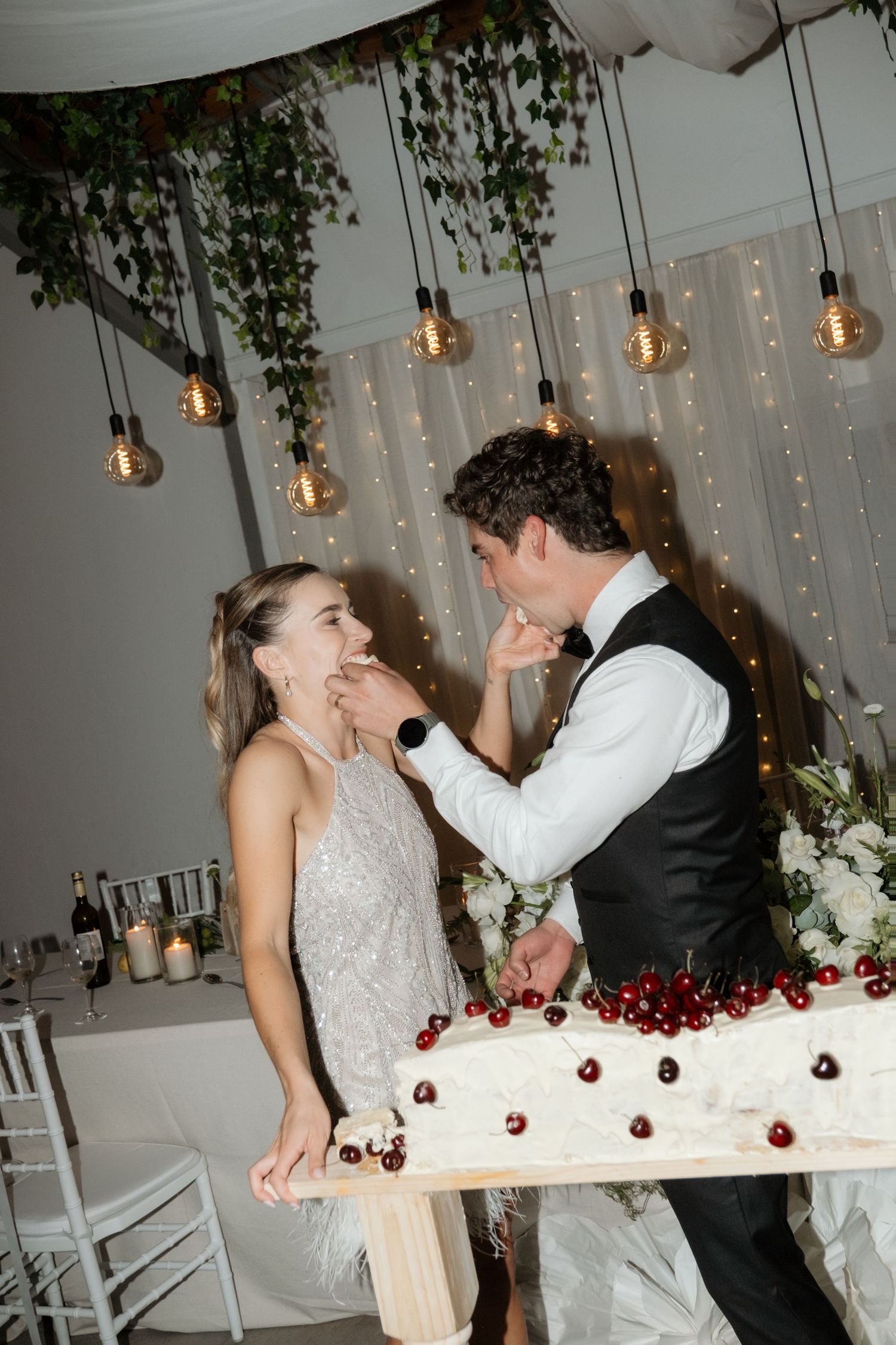 Groom feeding wedding cake to bride in sequin dress at decorated reception with cherry cake.