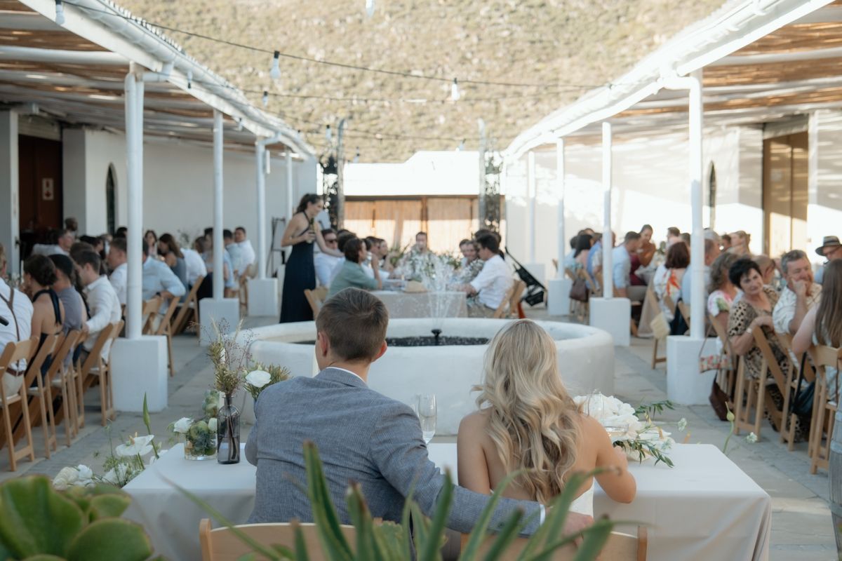 Elegant outdoor wedding reception with white decor, string lights, and guests seated at long tables under a pergola.
