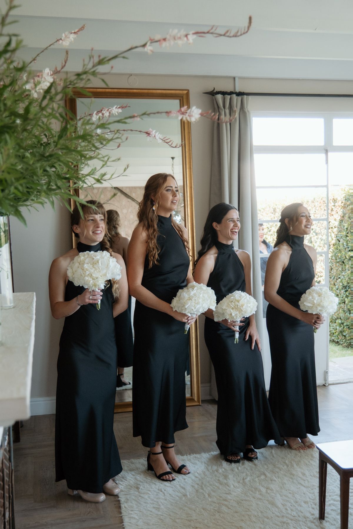 Four bridesmaids in matching black halter gowns holding white hydrangea bouquets indoors.