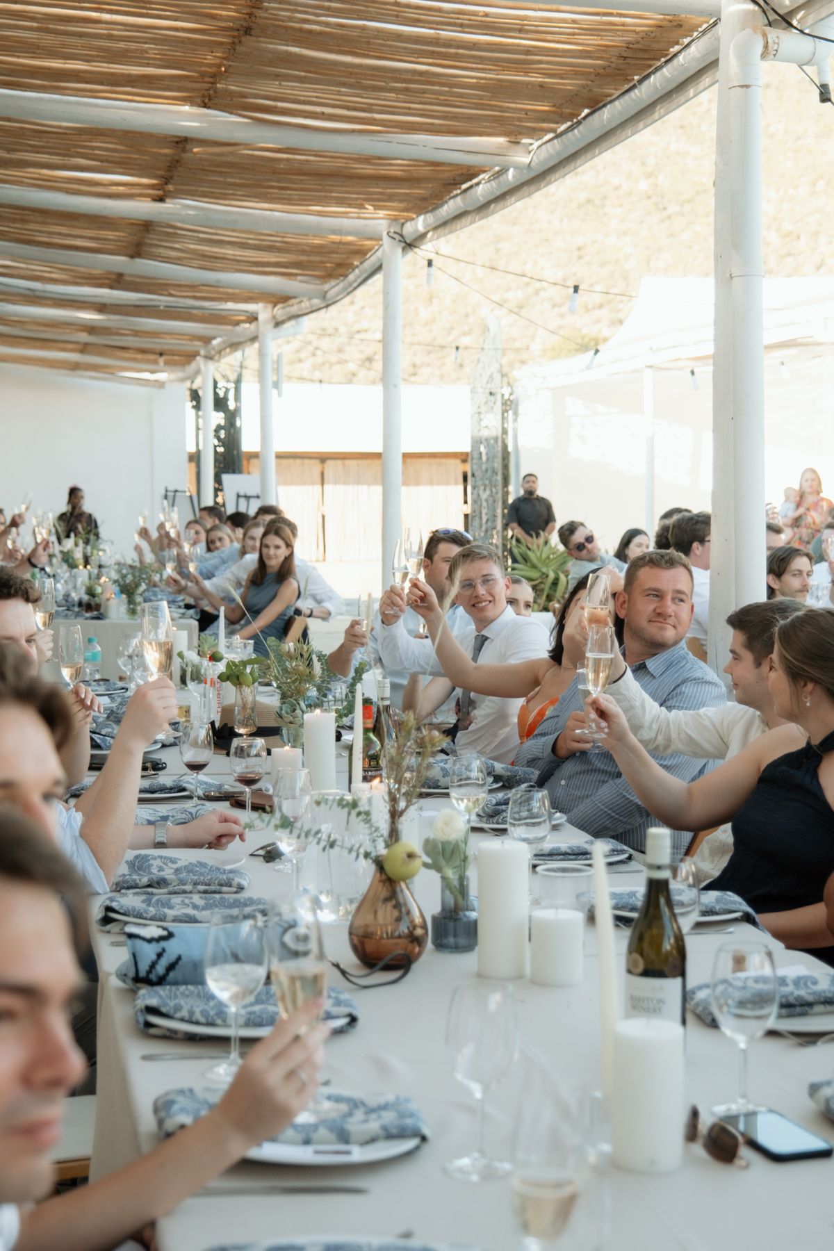 Wedding guests raising champagne glasses in a toast at a long decorated reception table under a rustic reed ceiling.