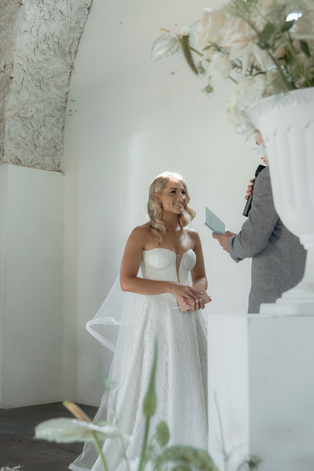 Smiling bride in strapless white lace gown during wedding ceremony beside floral arrangement.