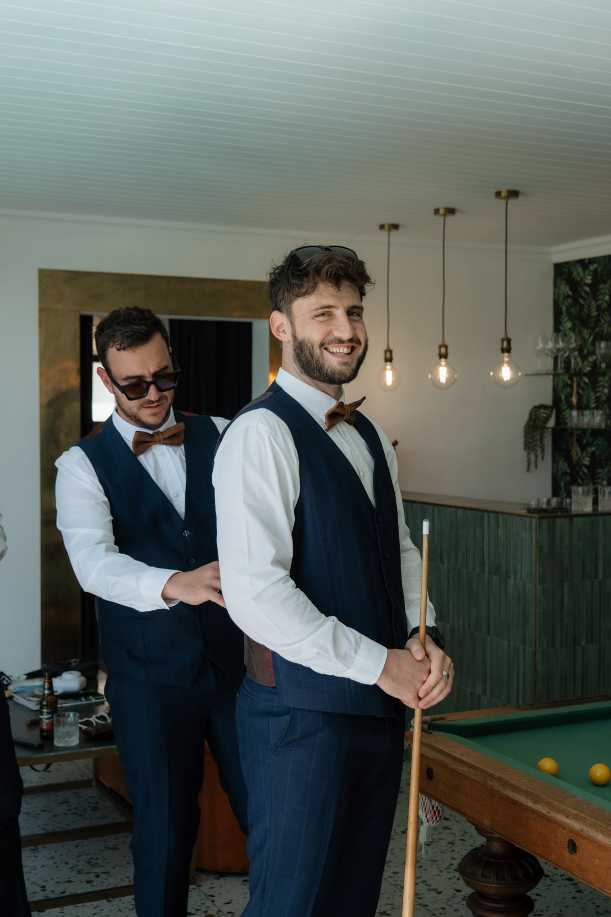 Two groomsmen in navy vests and bow ties smile near a billiard table indoors.