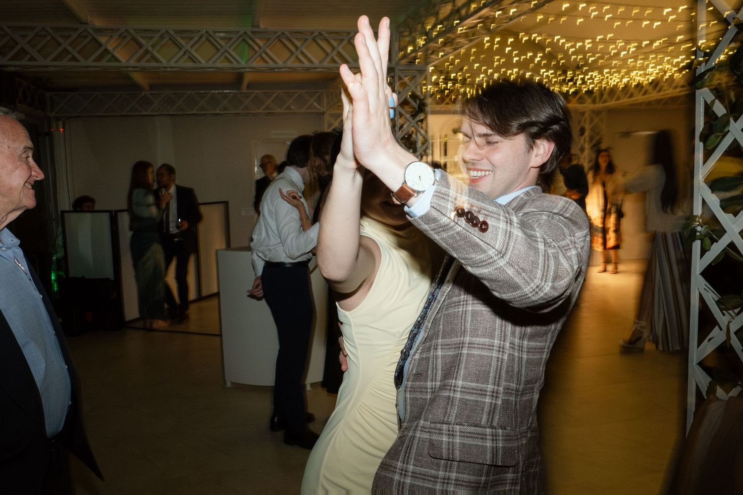 Couple high-fiving and dancing joyfully at wedding reception under sparkling fairy lights.