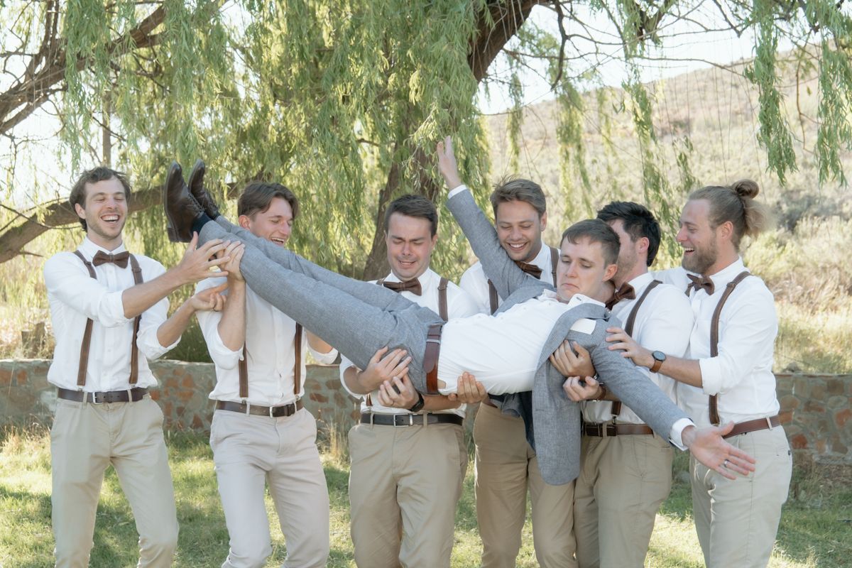 Groomsmen in suspenders playfully lifting groom outdoors under willow tree before wedding ceremony.