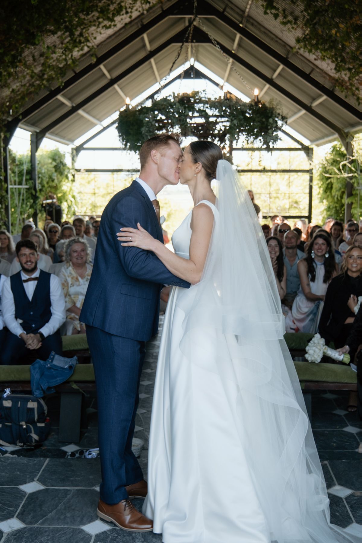 Bride and groom share first kiss at altar in glass greenhouse chapel surrounded by wedding guests.