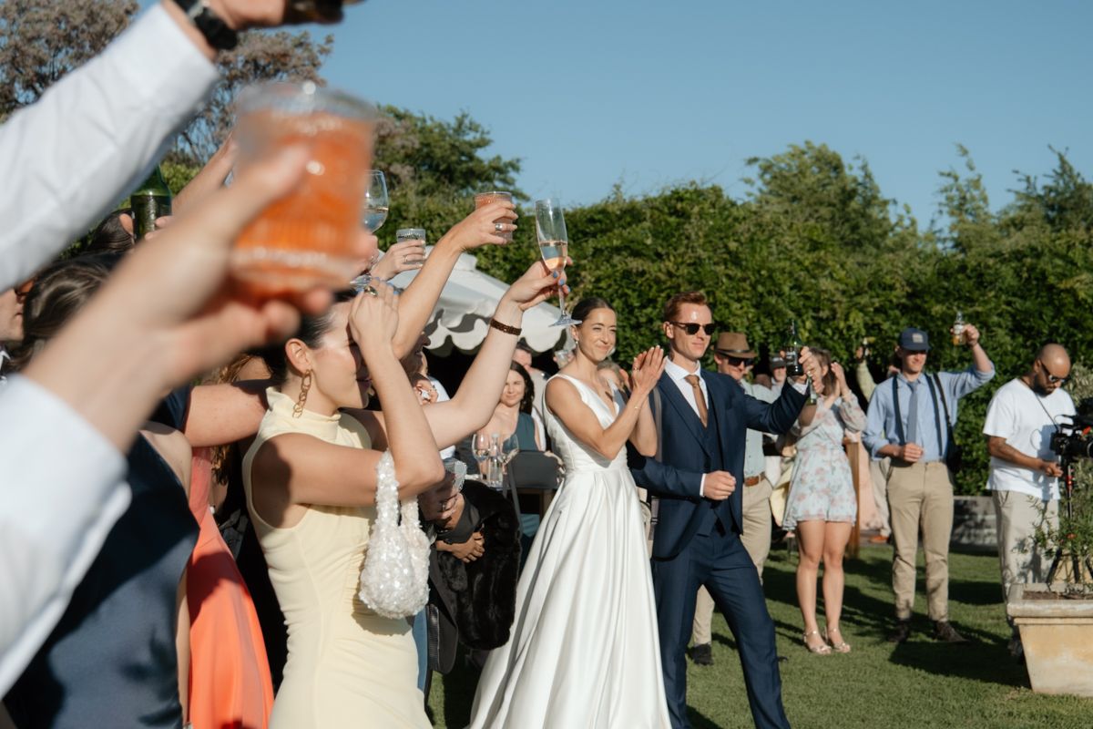 Newlyweds walk through celebratory guest tunnel raising glasses outdoors on bright sunny wedding day.
