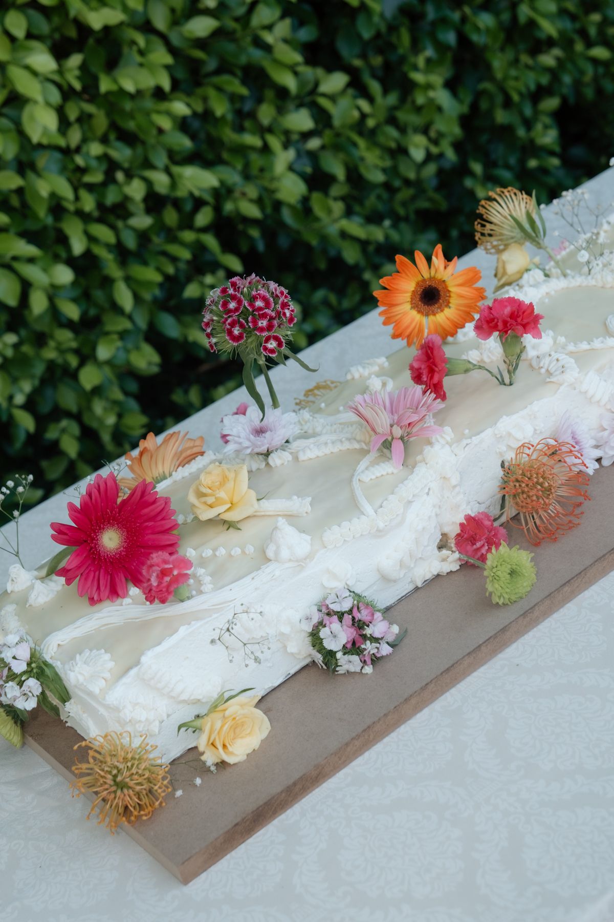 Elegant white wedding cake decorated with colorful fresh flowers on rustic wooden board outdoors.