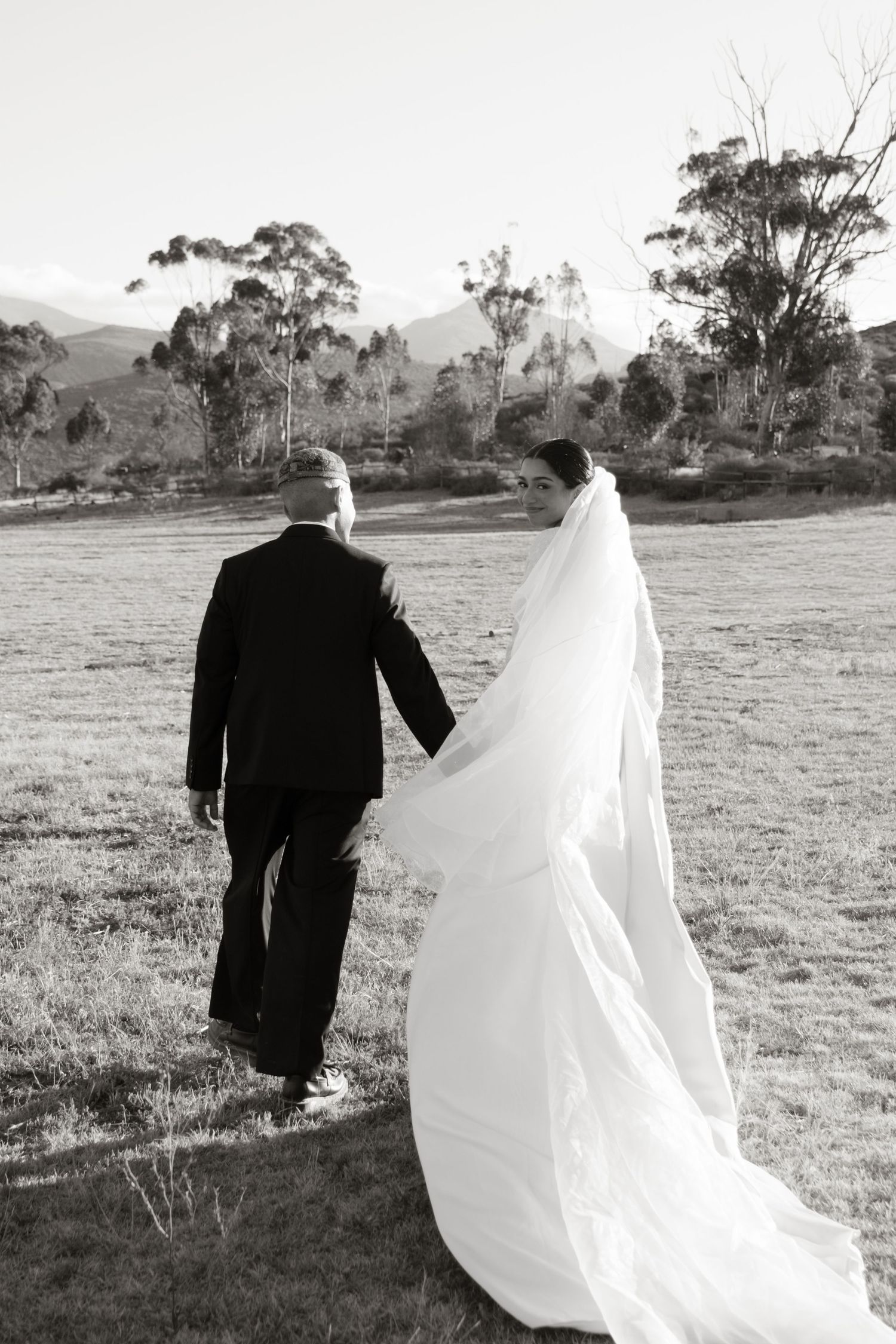Bride and groom walking hand in hand across a scenic field in black and white wedding photo.