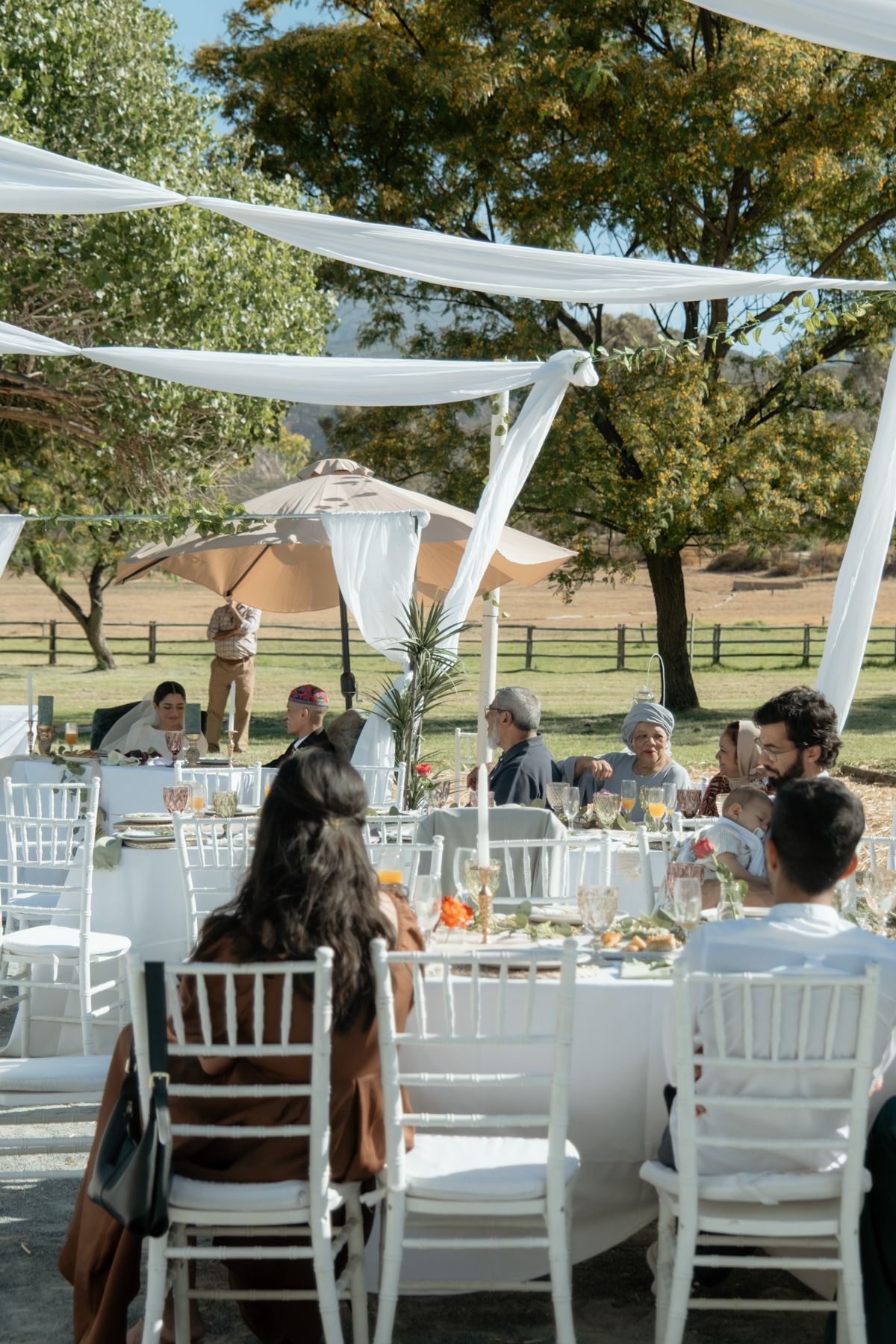 Guests seated at white round tables enjoy an outdoor wedding reception under draped fabric canopy.