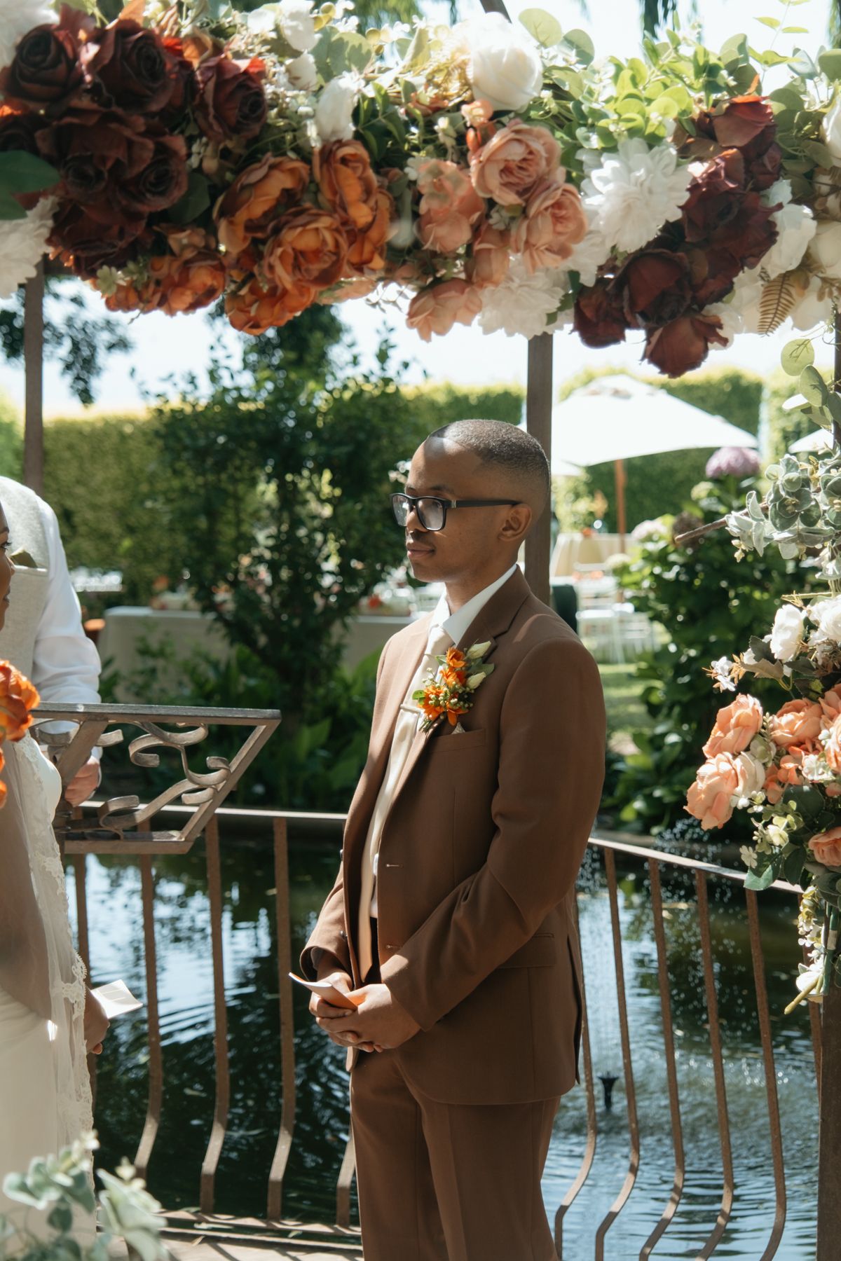 Groom in tan suit with floral boutonniere stands at altar beneath colorful rose wedding arch.