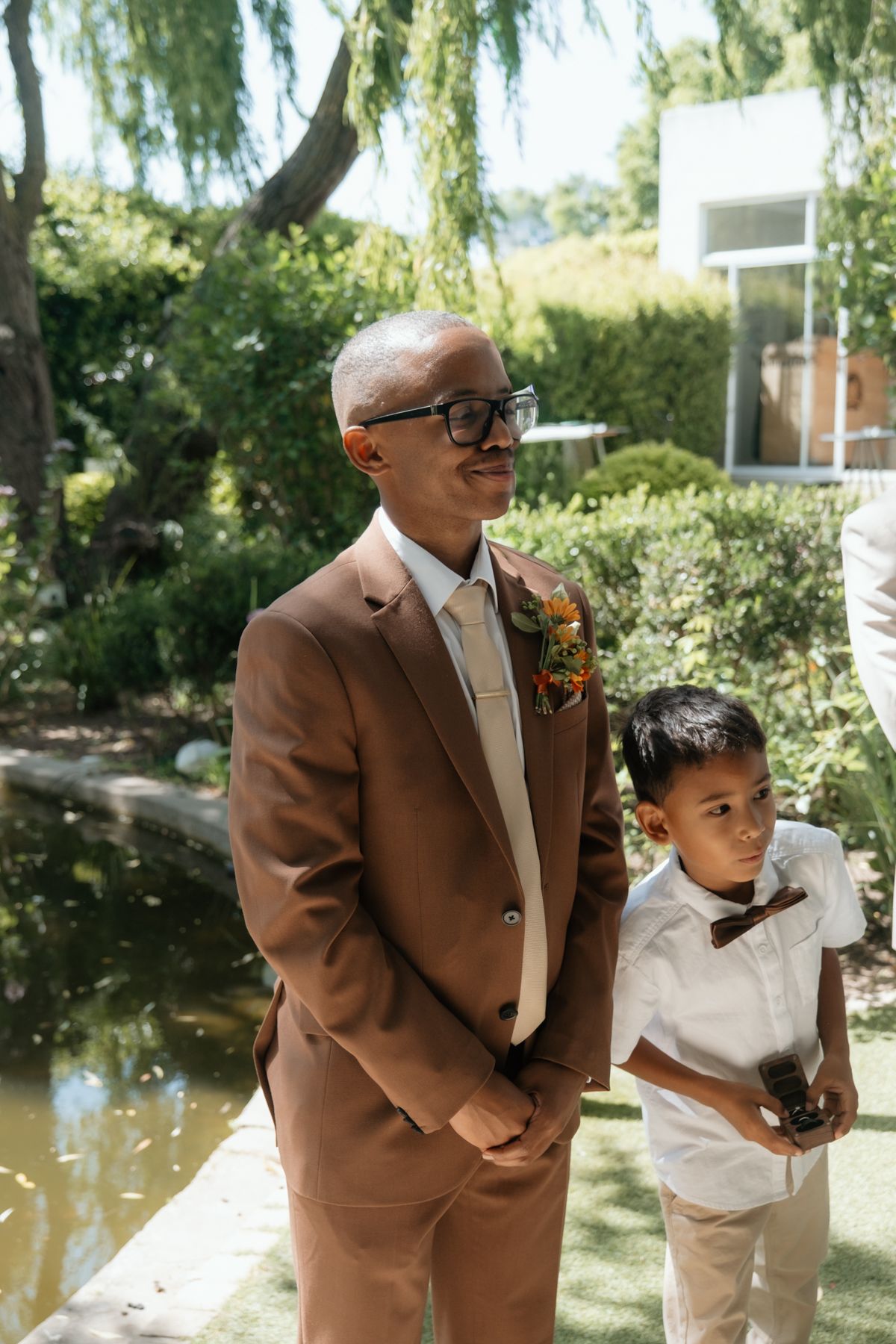 Groom in brown suit with boutonniere stands beside young ring bearer in white shirt at outdoor wedding ceremony.