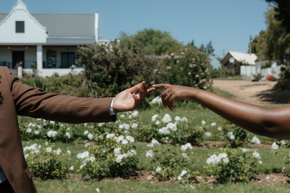 Two people gently touching fingertips in a garden with white flowers and a white farmhouse in the background.