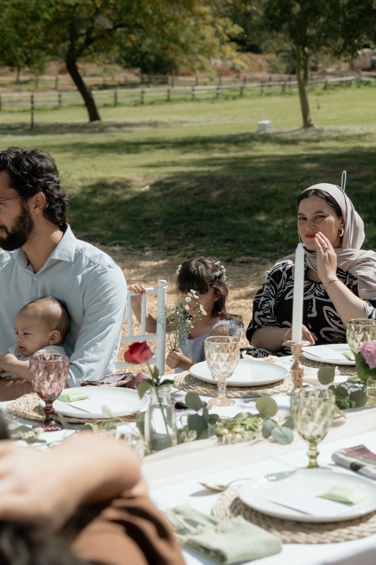 Guests seated at elegant outdoor wedding reception table in sunny garden setting.