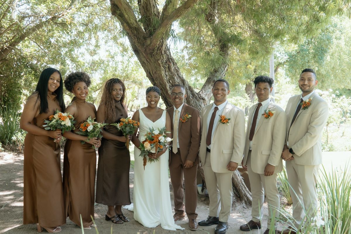 Wedding party poses under a tree, bridesmaids in brown dresses, groomsmen in tan suits, bride in white holding orange bouquet.