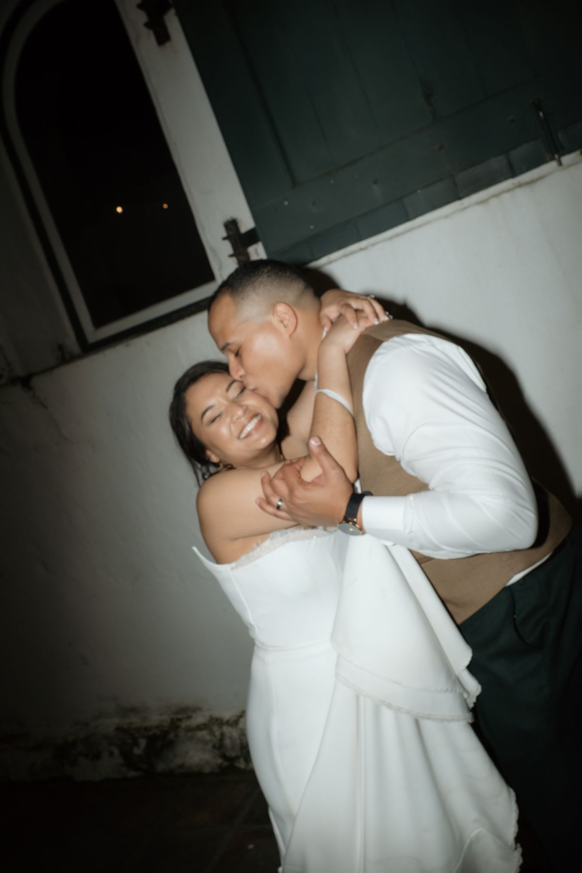 Groom kissing bride on cheek as she laughs at night.