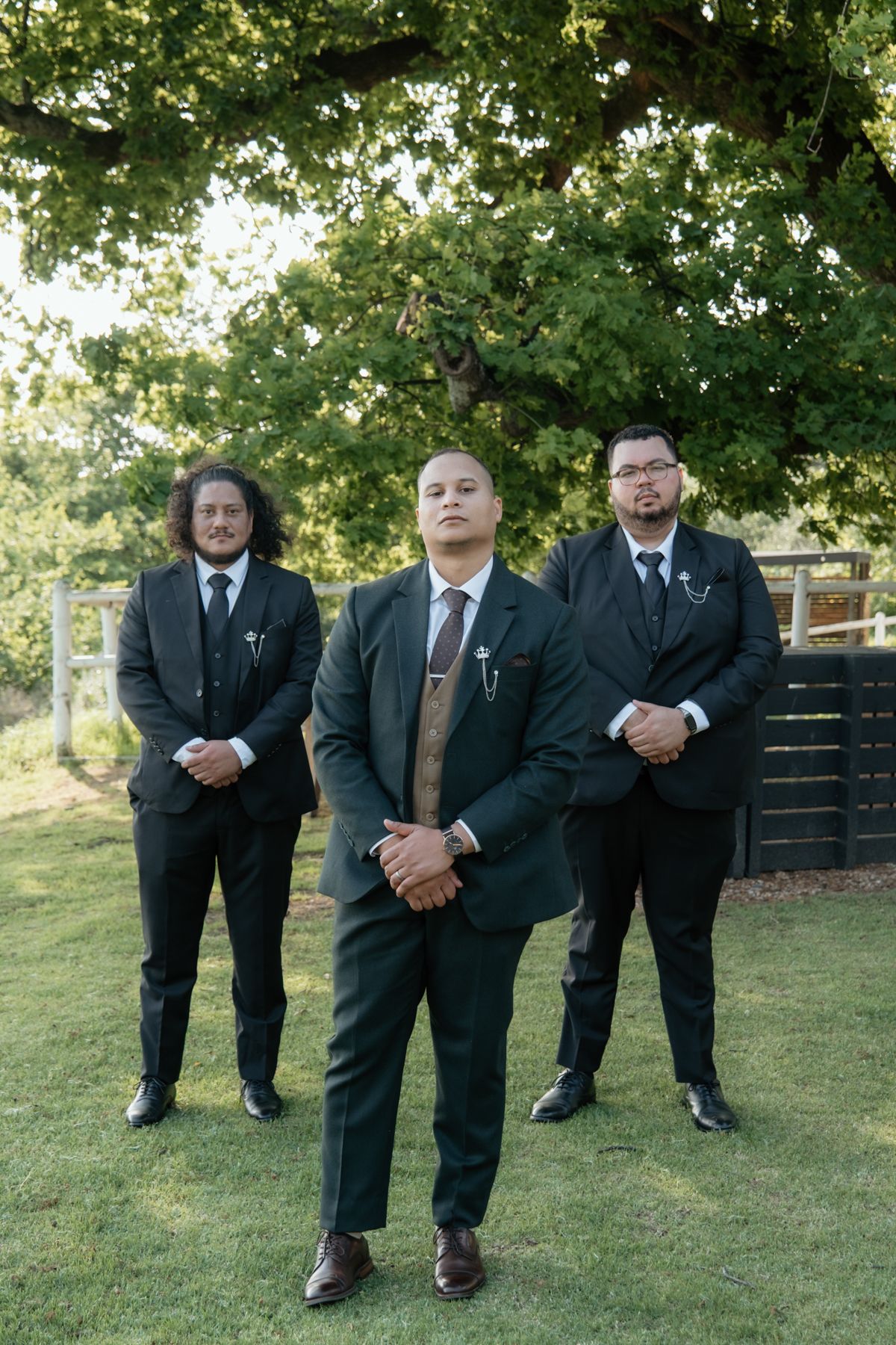 Groom and two groomsmen in dark suits posing seriously under a large oak tree on green lawn.