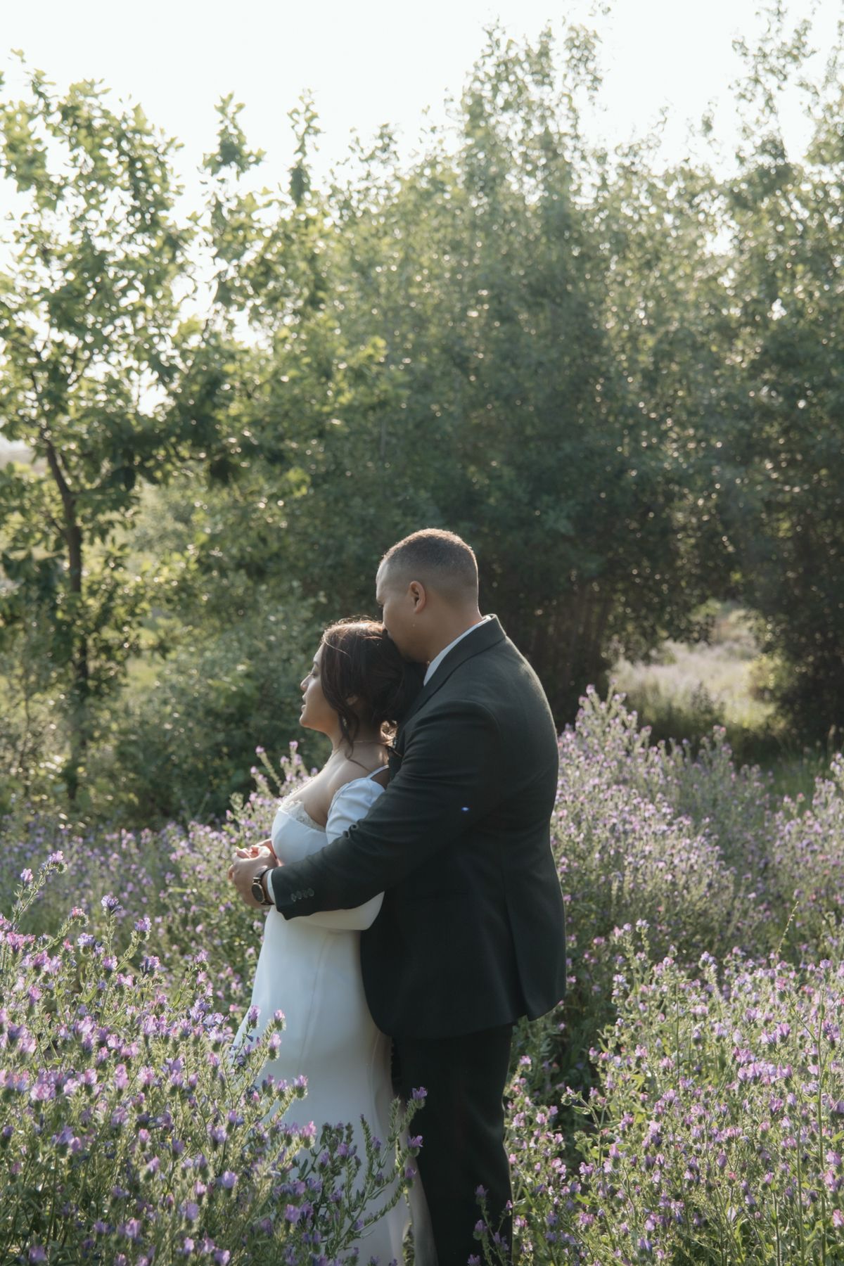 Couple embracing romantically in a lavender field surrounded by green trees at golden hour.