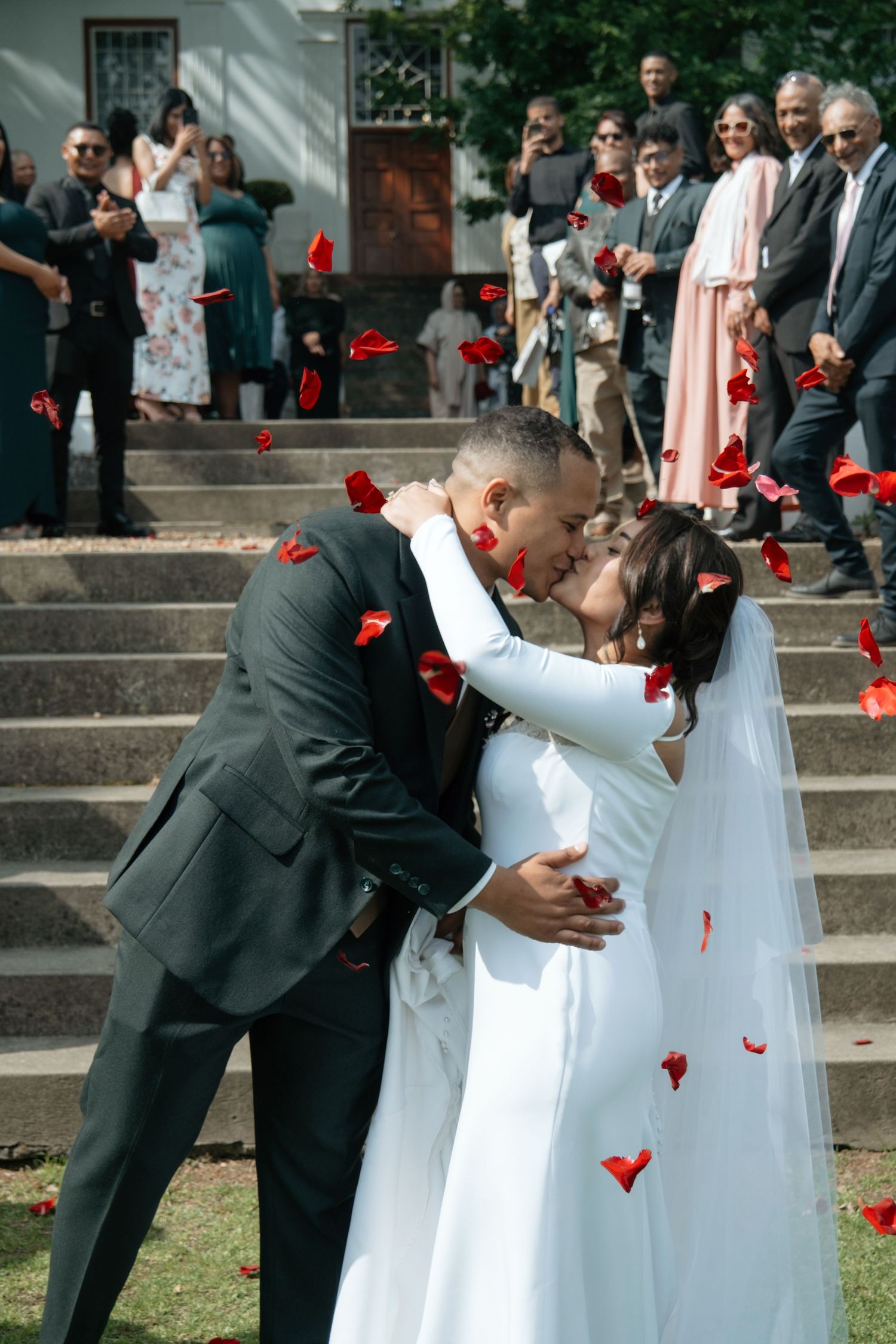 Bride and groom share a kiss as red rose petals fall around them on church steps with wedding guests.