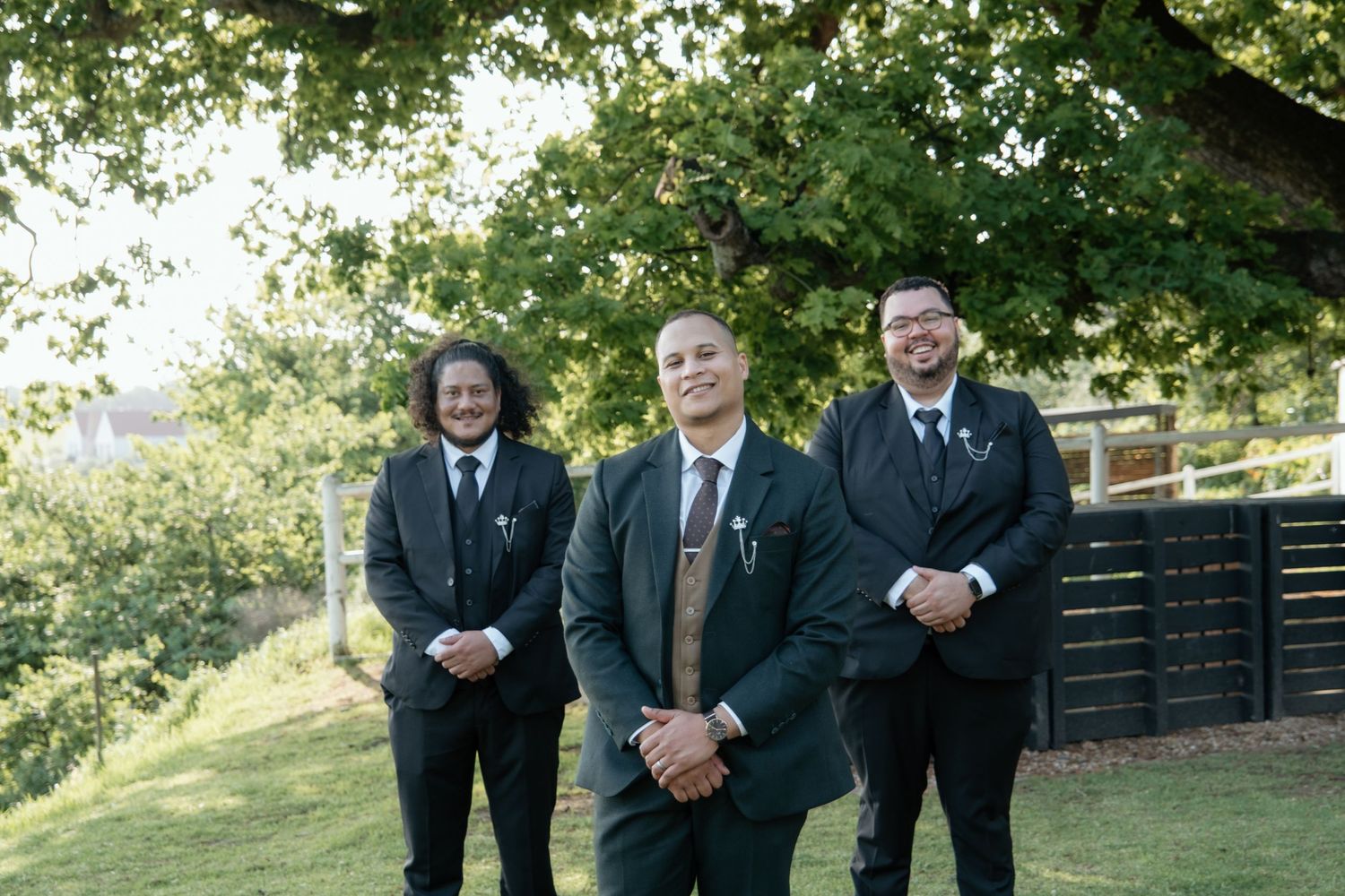 Three groomsmen in dark suits stand outdoors on green grass beneath a large tree at a wedding.