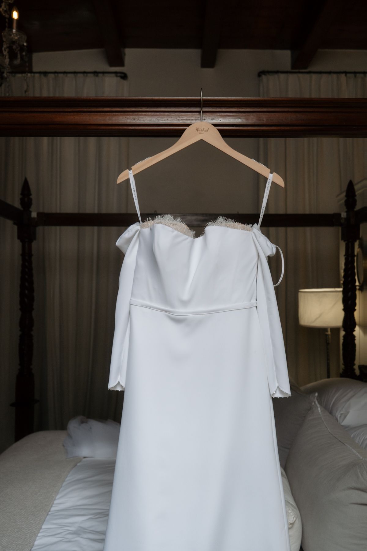 Elegant white off-shoulder wedding dress with long sleeves hanging on a wooden hanger above a four-poster bed.