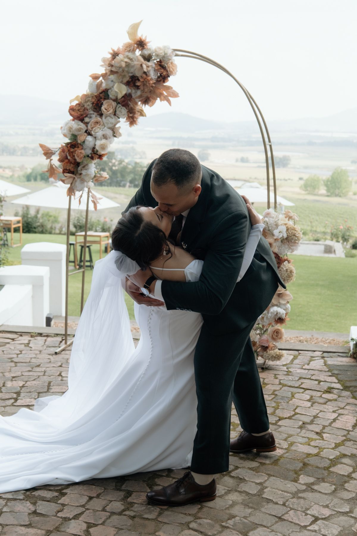 Bride and groom sharing first kiss under floral circular arch at outdoor wedding ceremony.