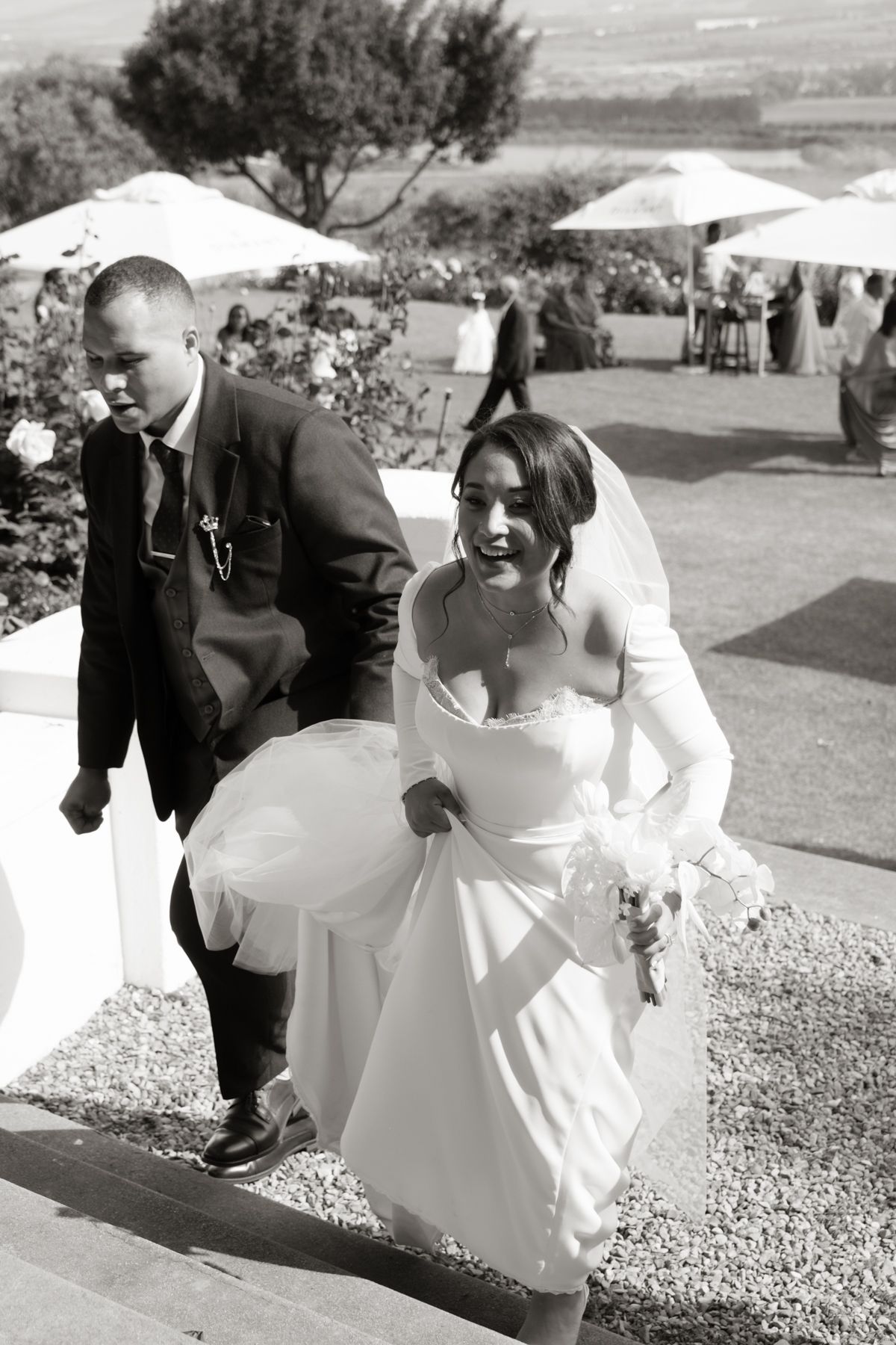 Black and white photo of smiling bride in white gown and veil walking with groom at outdoor wedding.