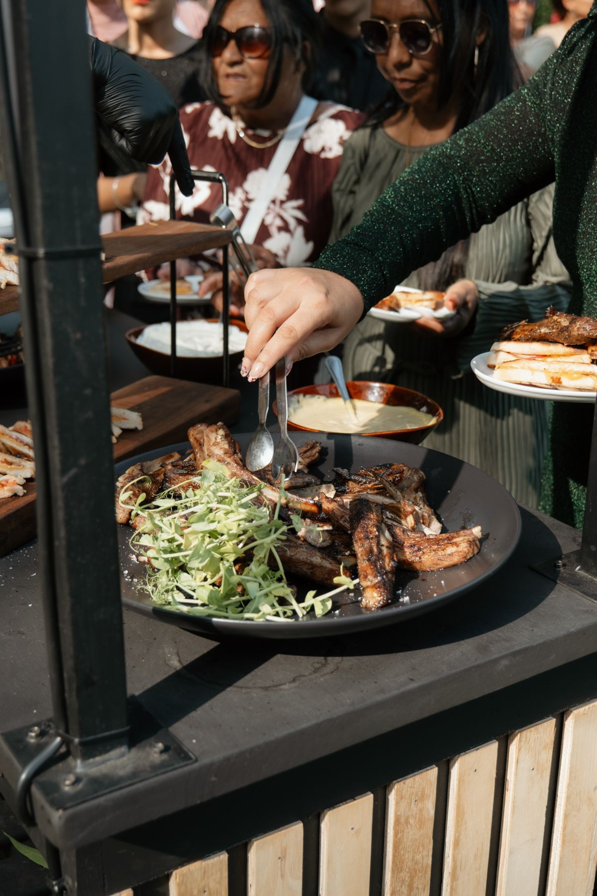 Wedding guests serving themselves grilled meat and greens at outdoor buffet reception station.