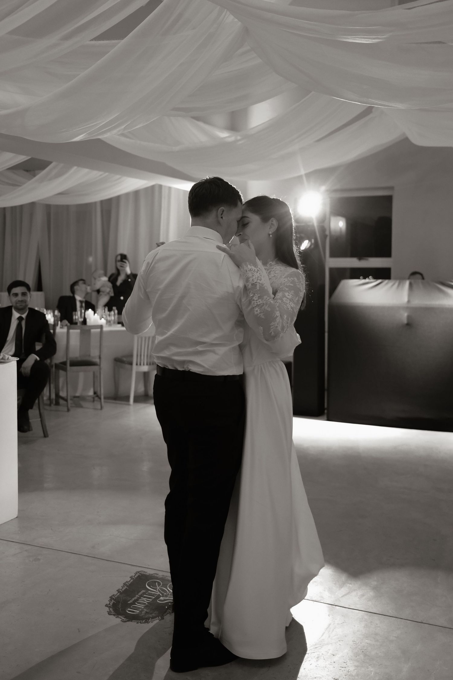 Black and white photo of bride and groom sharing first dance under draped ceiling at wedding reception.