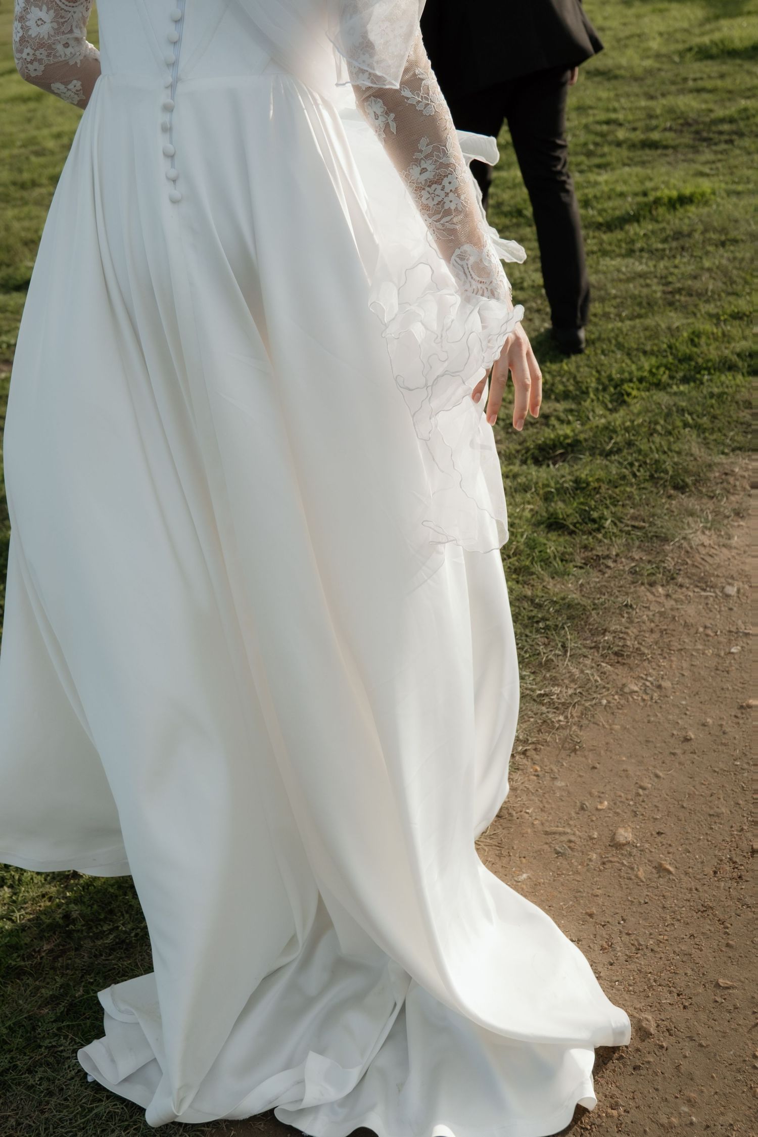 Bride walking in elegant white lace long-sleeve wedding gown with flowing train on outdoor path.