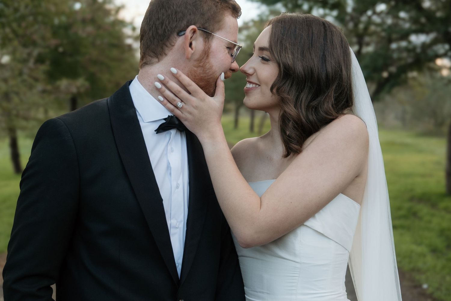 A romantic wedding couple shares an intimate moment outdoors with the bride in white and groom in black tuxedo.
