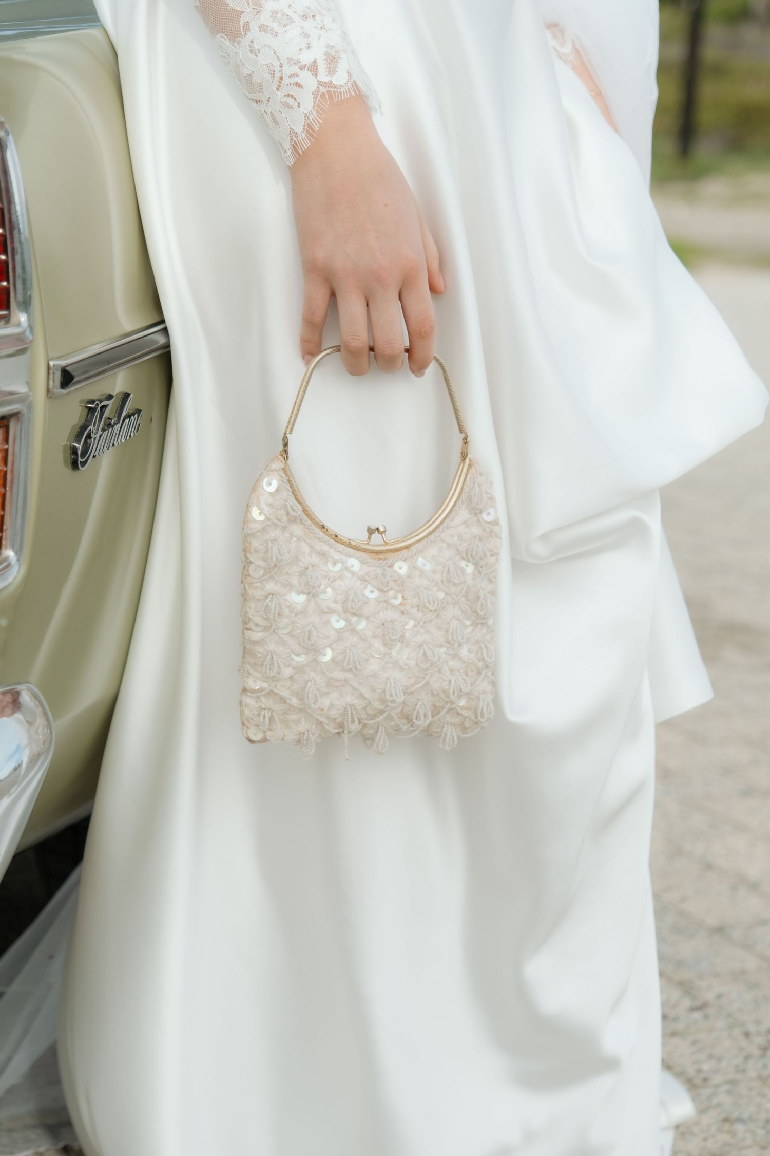 Bride in white gown holding a sequined ivory clutch purse with gold handle beside a vintage car.