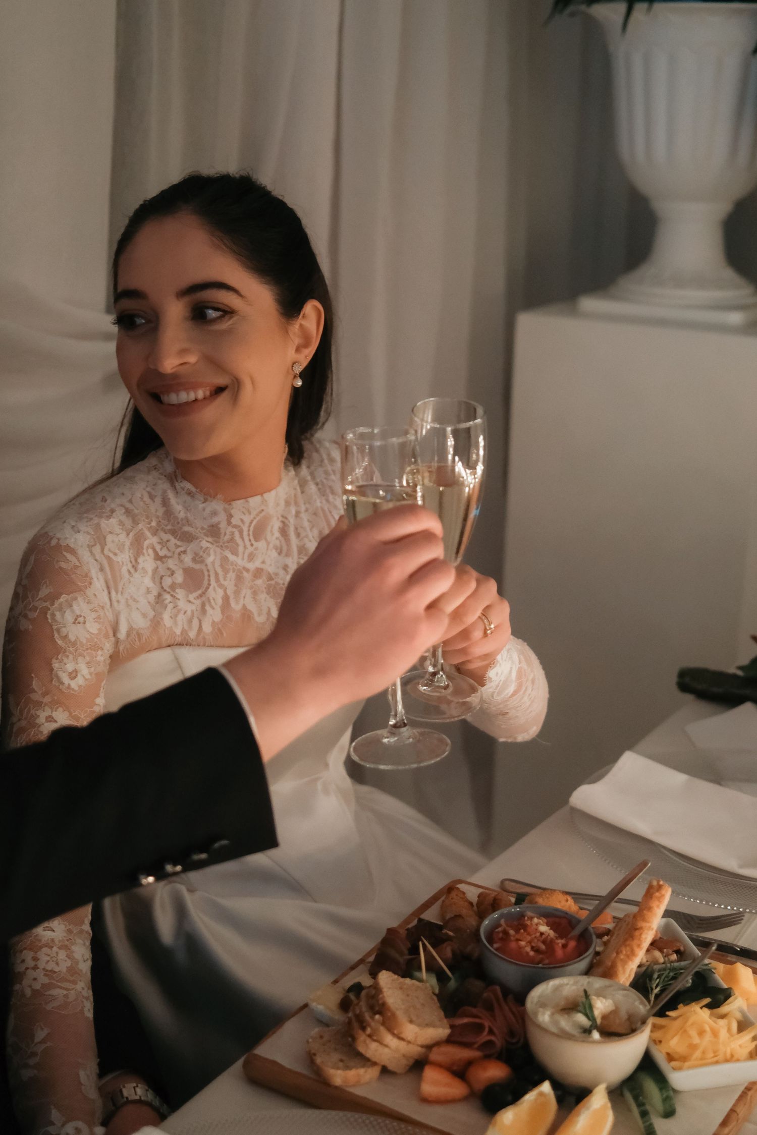 Smiling bride in lace gown clinks champagne glasses at wedding reception table with food spread.