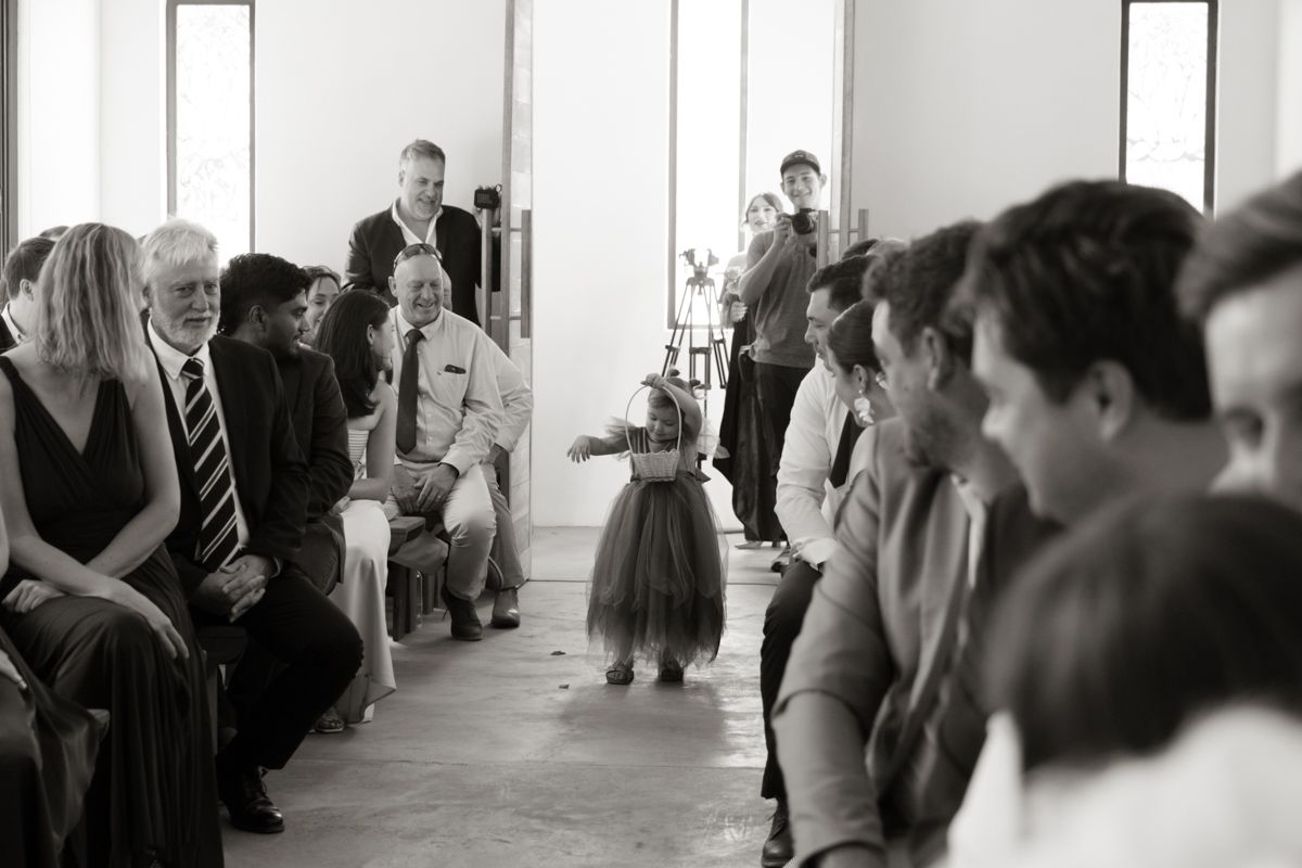 Flower girl in tutu dress walks down wedding aisle as guests watch during indoor ceremony.