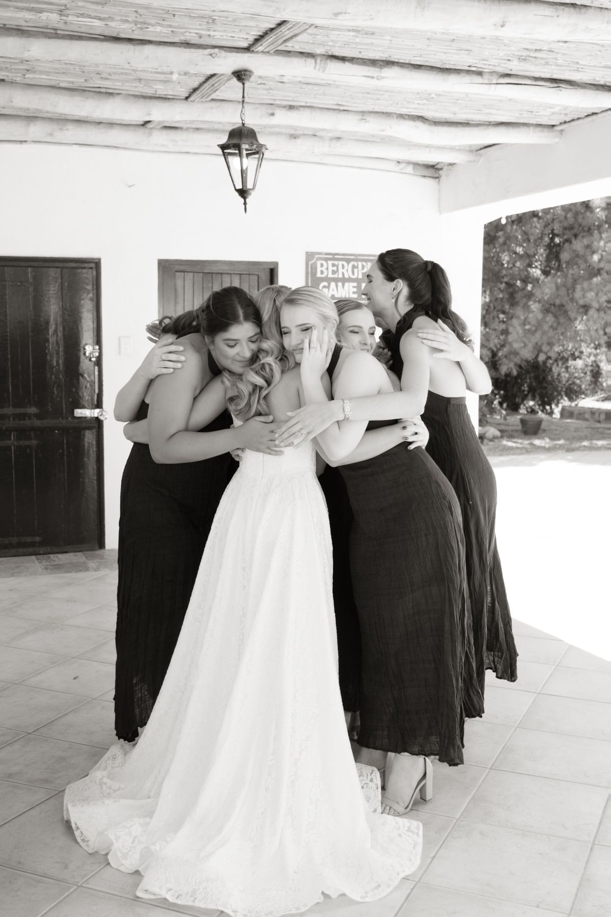 Black and white photo of bride embraced by four bridesmaids in group hug at wedding venue.
