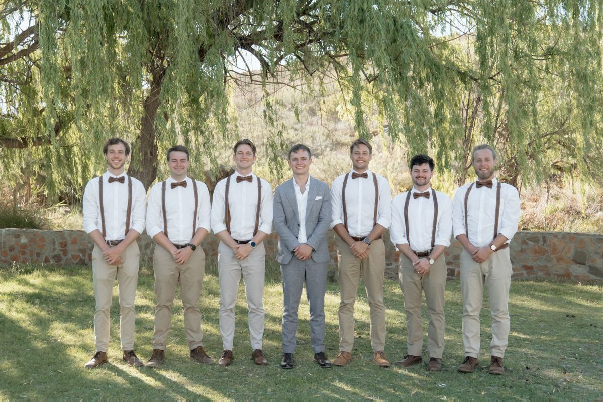 Groom in grey suit poses with groomsmen in khaki pants and suspenders under a willow tree.