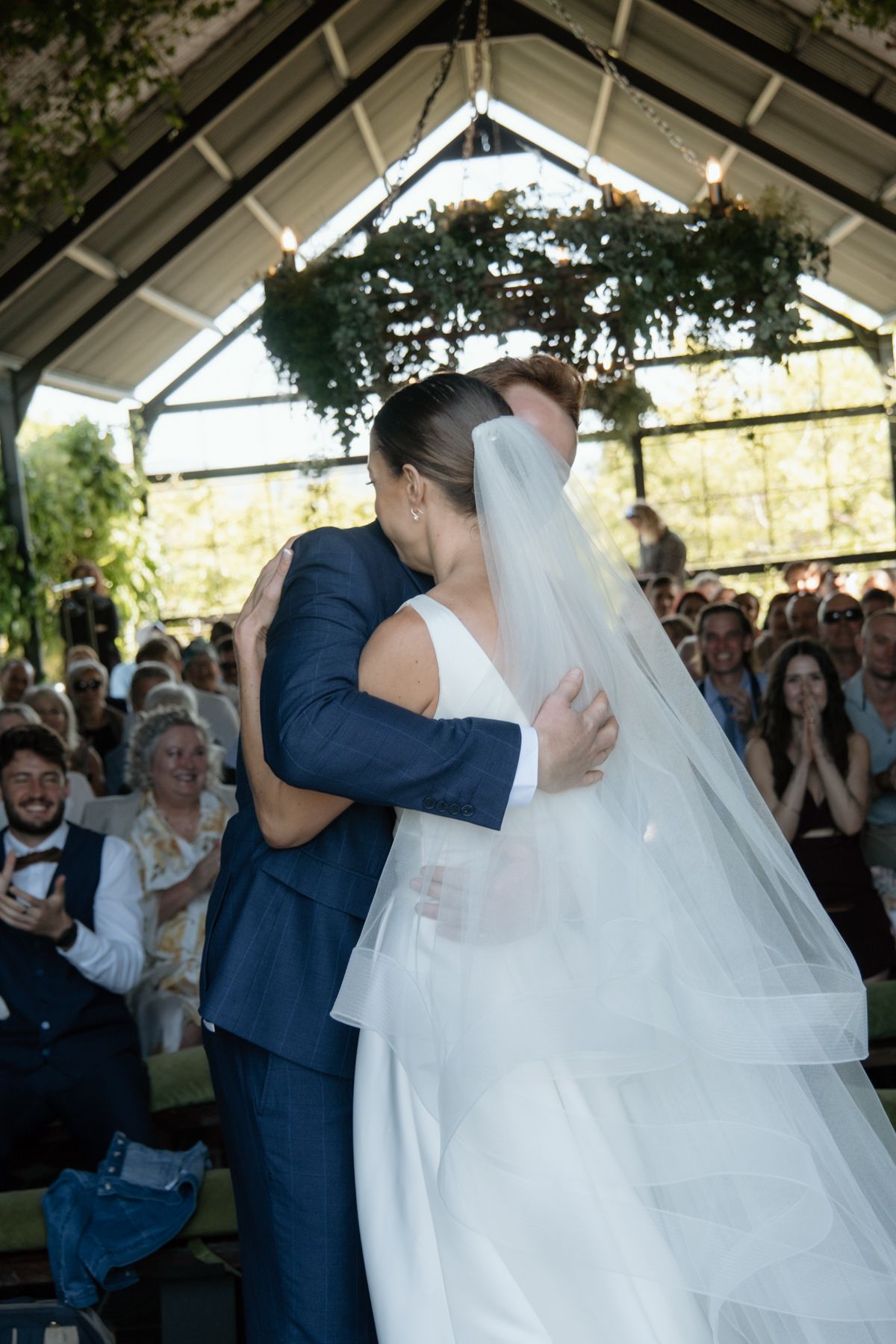 Bride and groom share their first kiss at the wedding altar, surrounded by applauding guests in a greenhouse venue.