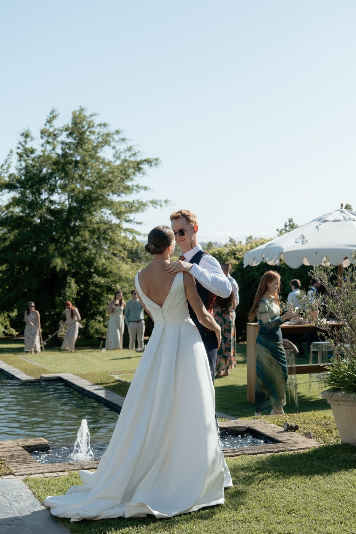 Bride in a backless white gown and groom share a romantic moment beside a garden pool with fountains.