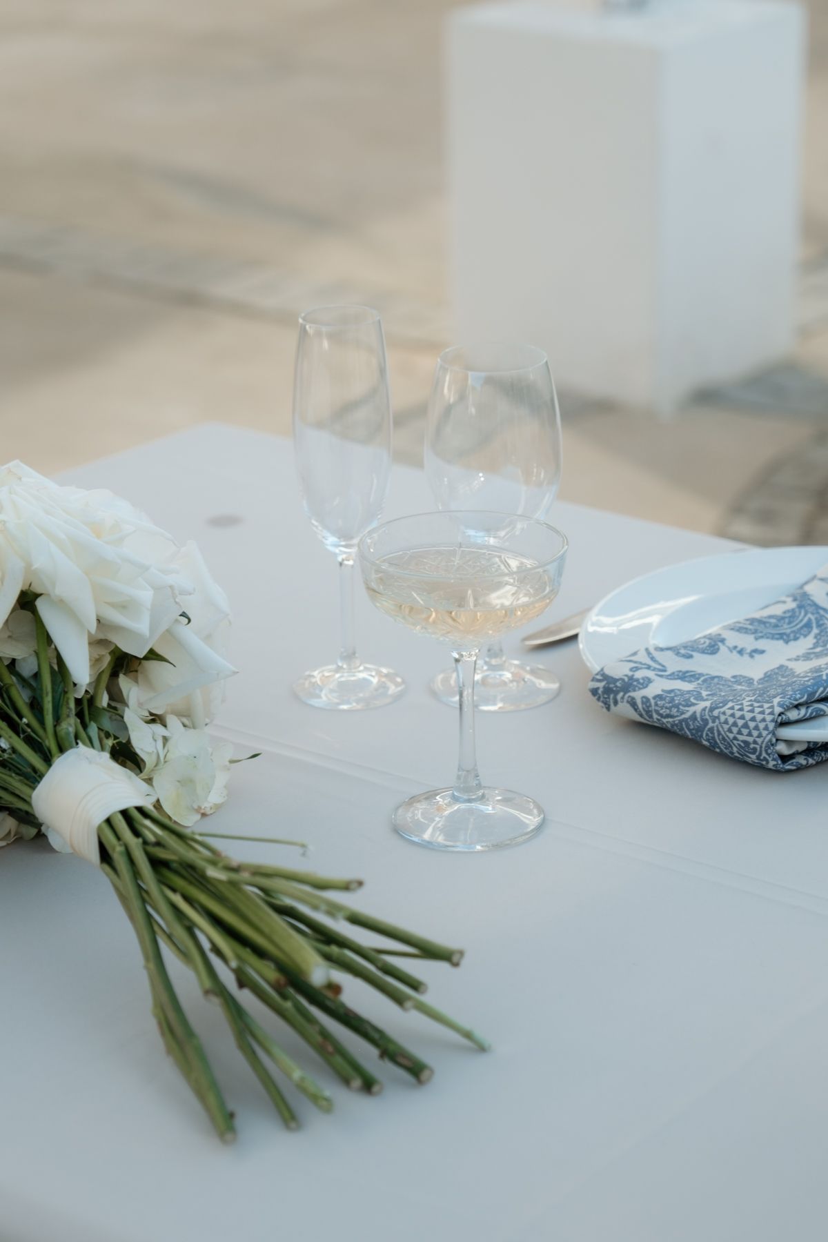 White bridal bouquet beside champagne glasses and blue napkin on white tablecloth at elegant wedding reception.