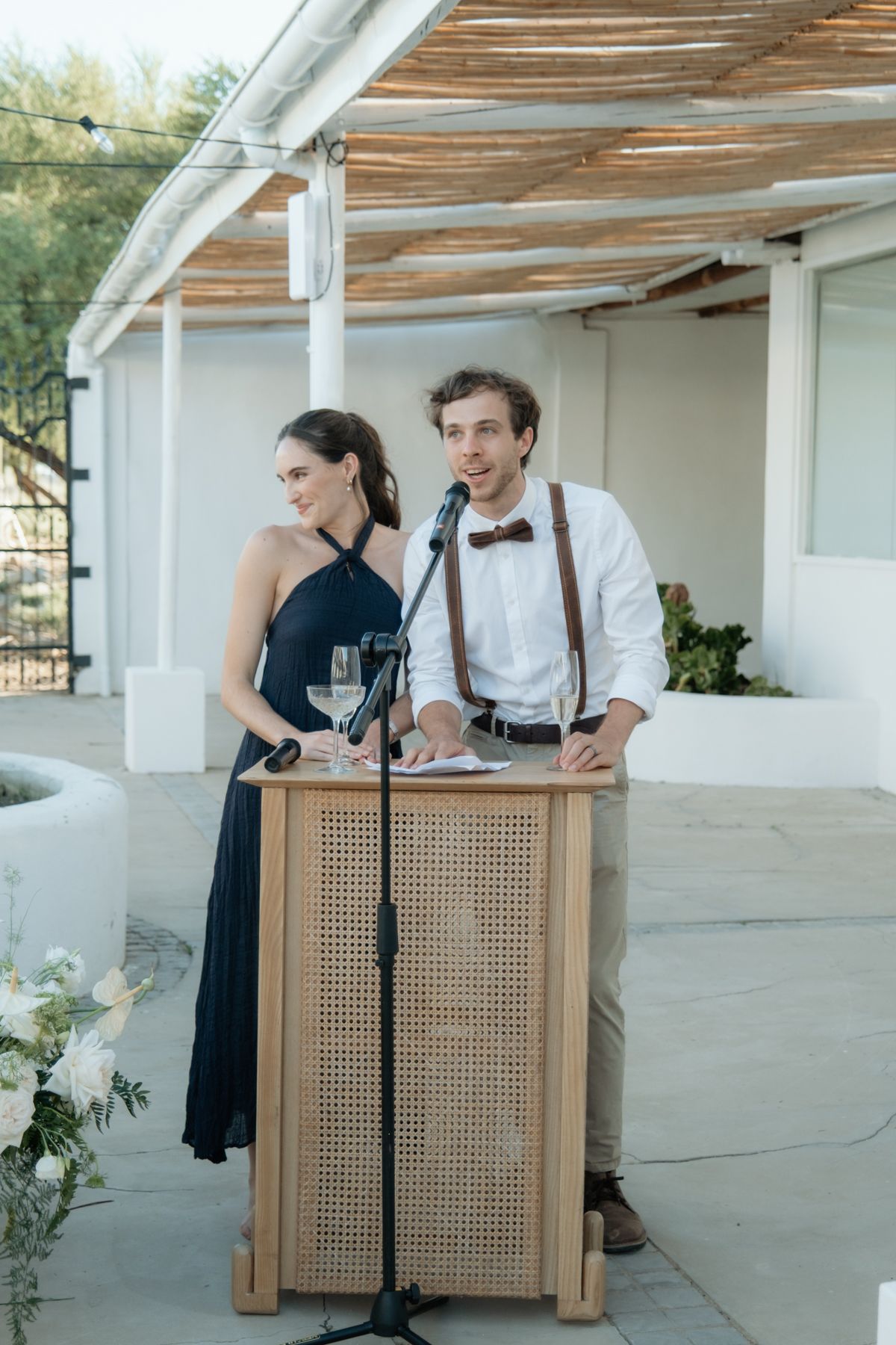 A man in suspenders and bow tie stands with a woman in a navy dress at a wooden podium during an outdoor wedding speech.