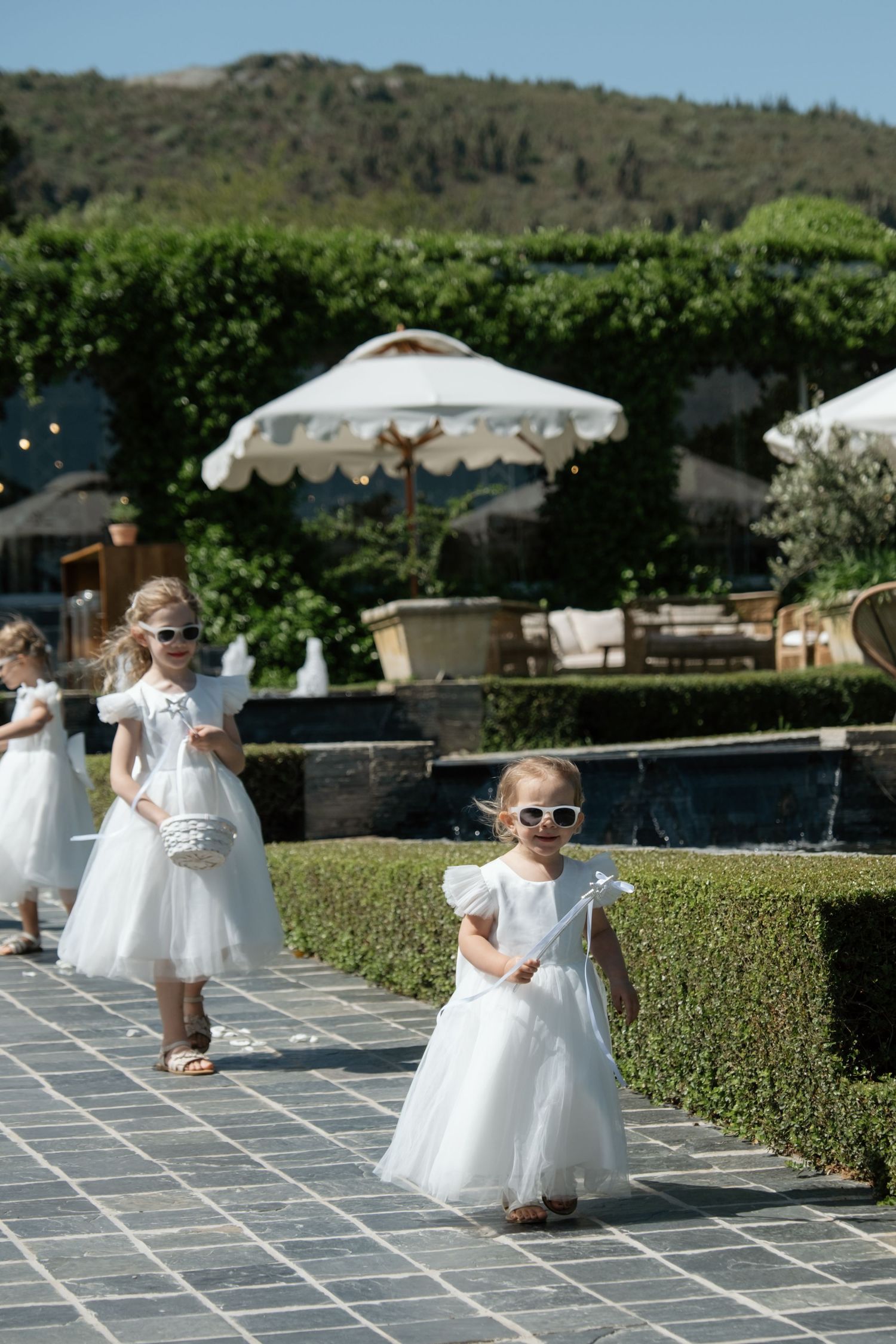 Adorable flower girls in white dresses and sunglasses walk along a garden path at an outdoor wedding.