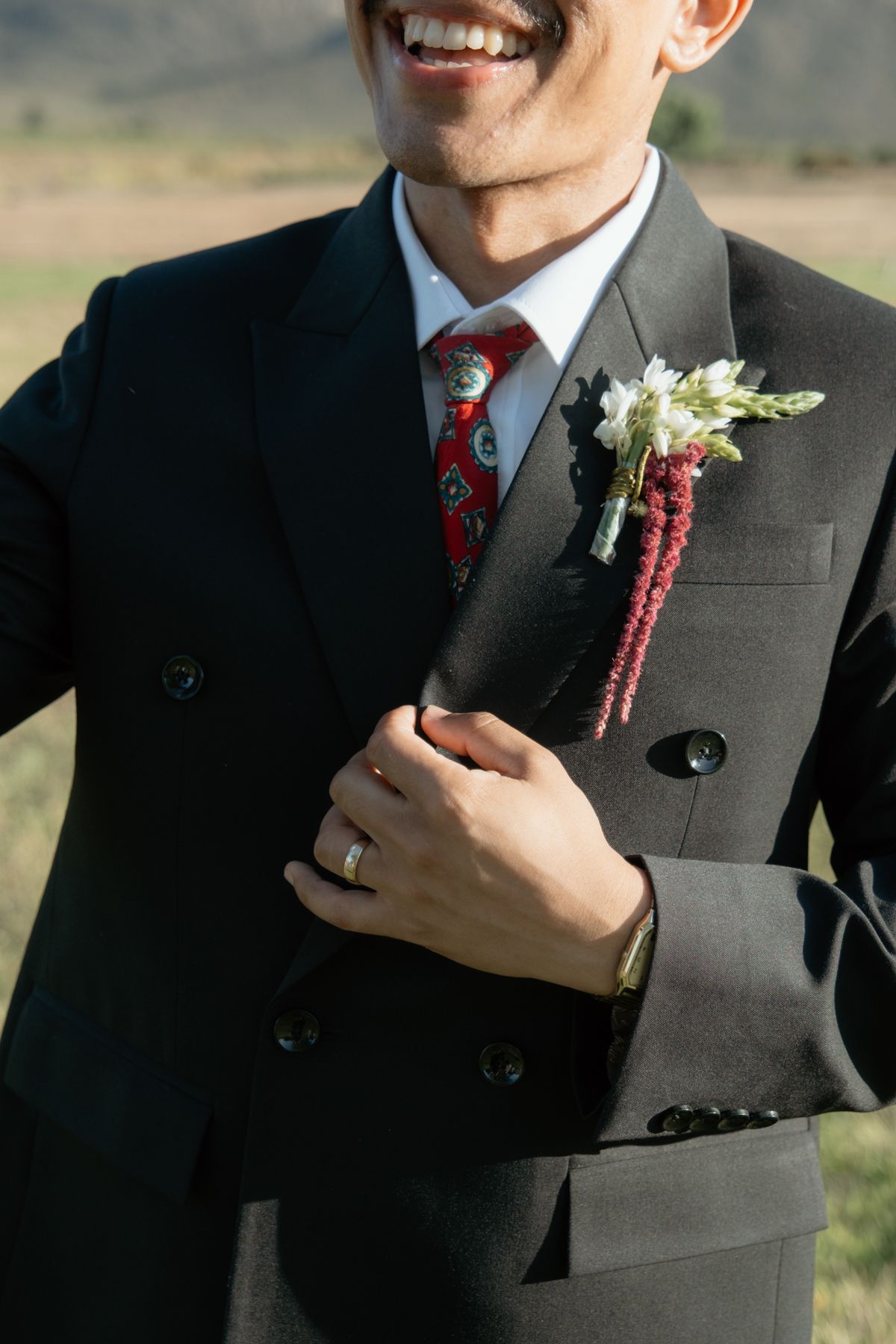Smiling groom in dark suit with red patterned tie and floral boutonniere, adjusting jacket lapel outdoors.