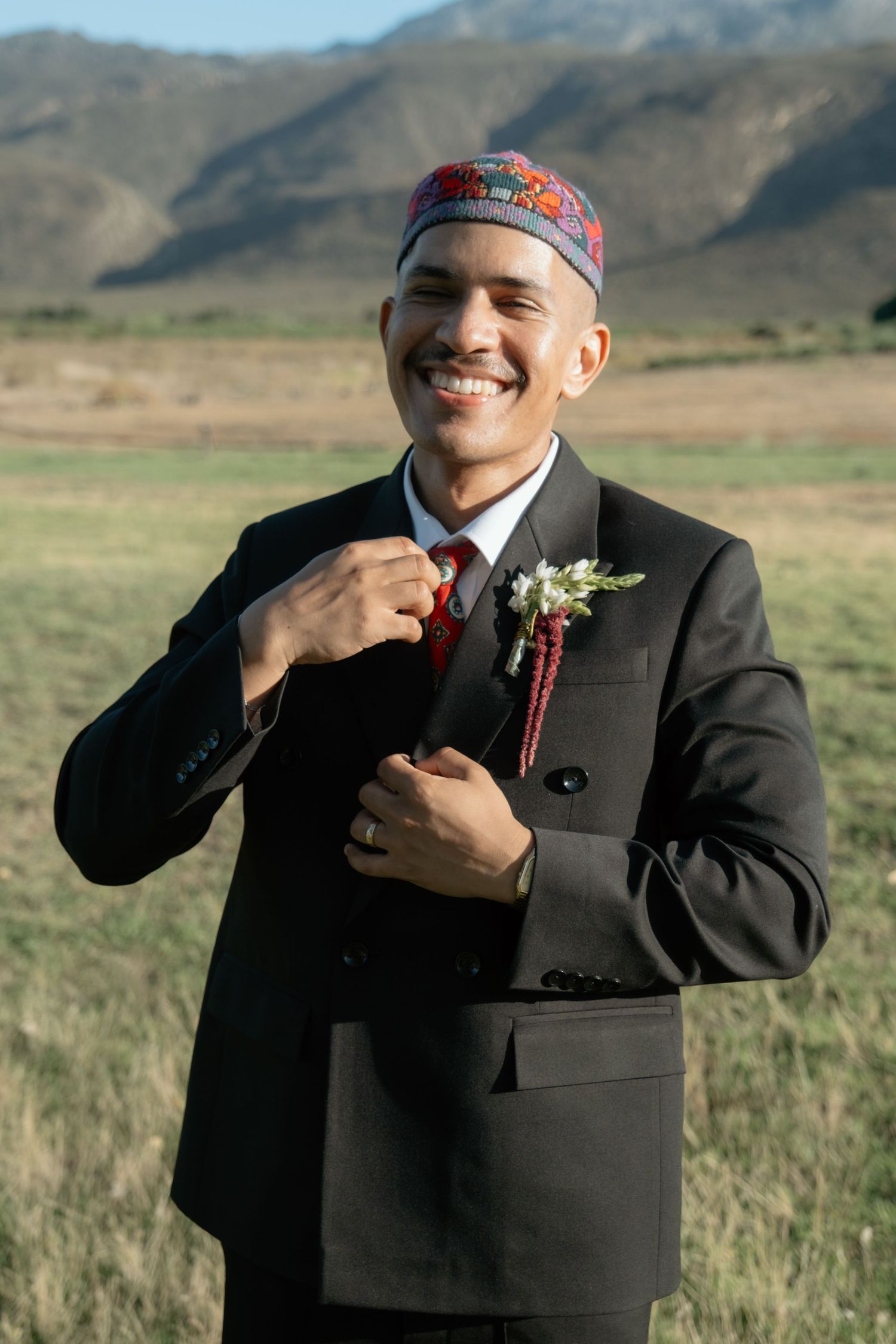 Smiling groom in black suit adjusts red tie wearing floral boutonniere and traditional kufi cap outdoors.