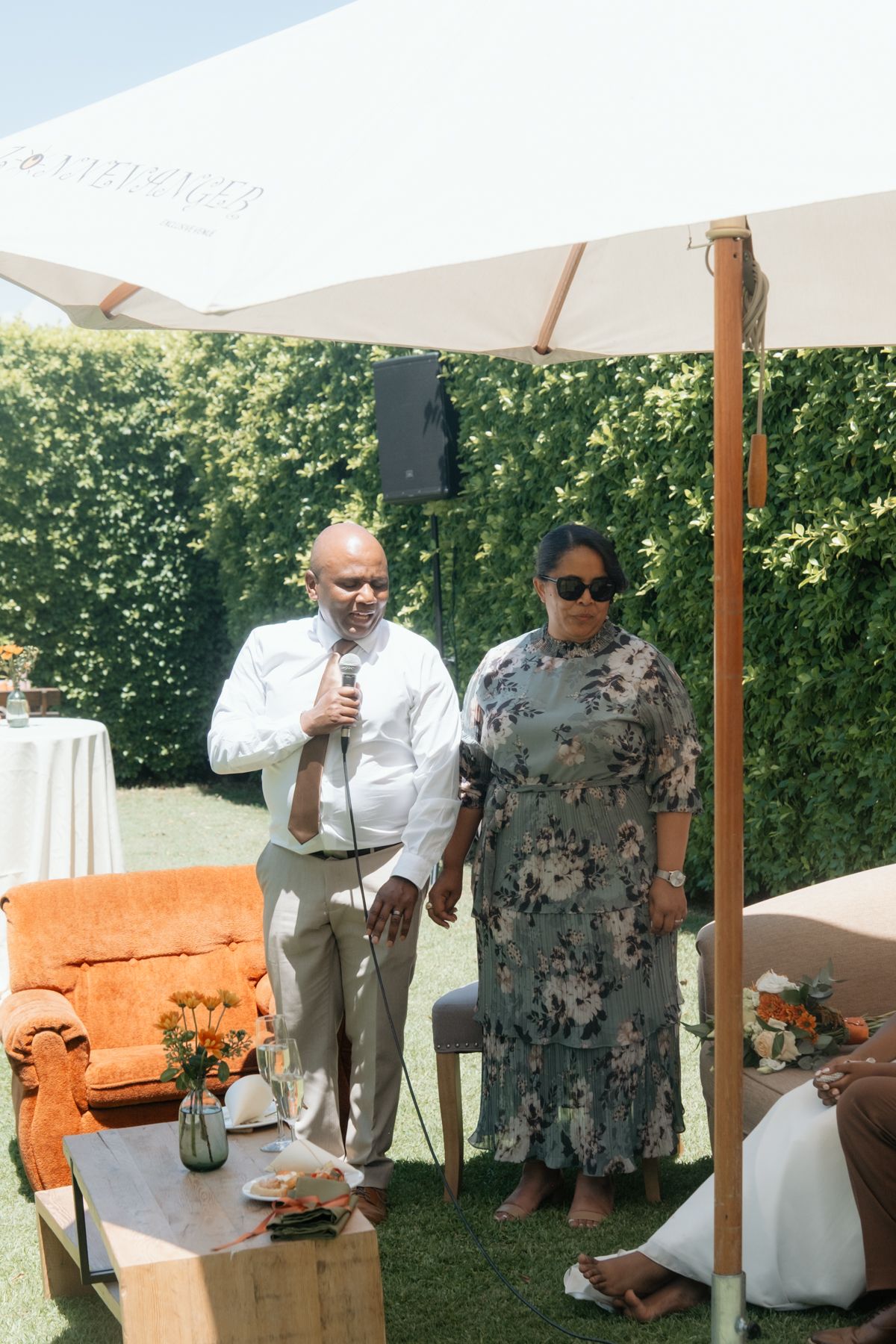 Couple speaks into microphone at outdoor garden party under white patio umbrella on sunny day.