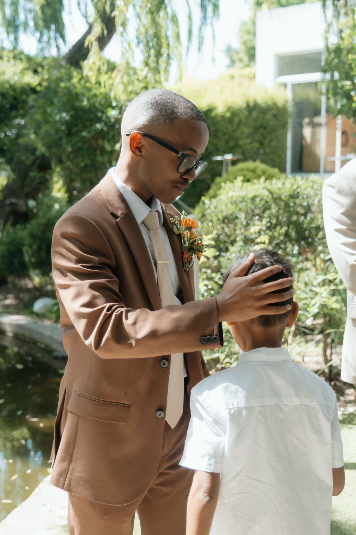 Groom in brown suit with floral boutonniere tenderly fixes young boy's hair at outdoor wedding ceremony.